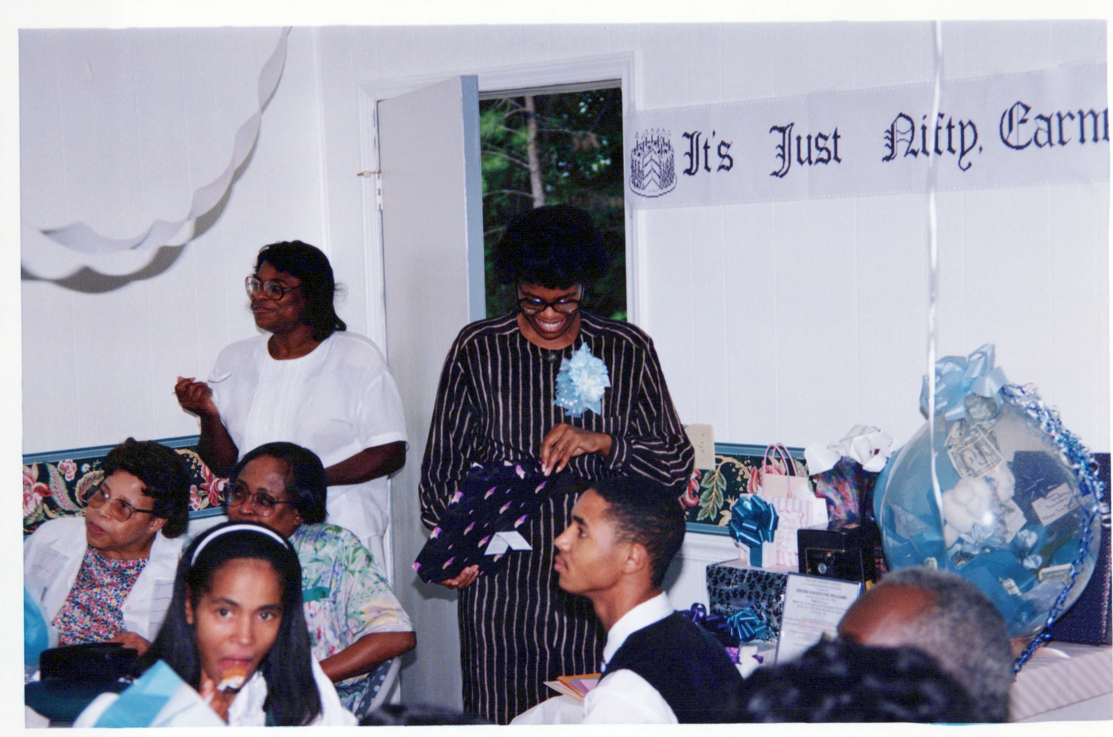 A smiling Black woman in a striped blouse and corsage opens a gift at her party, surrounded by friends and family. A birthday banner hangs on the wall.