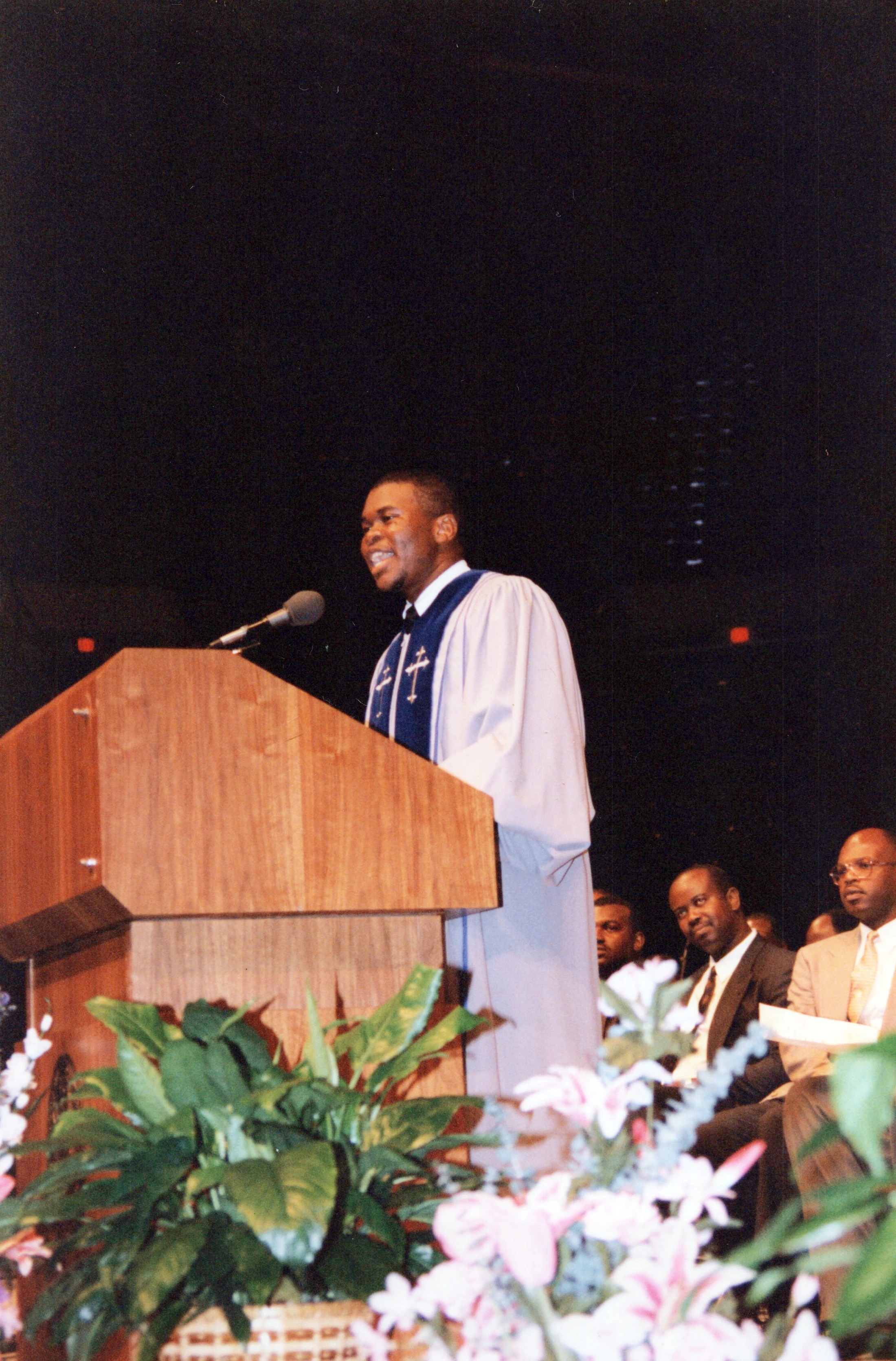 A young Black man in a light-colored robe and blue stole smiles as he speaks into a microphone at a wooden podium in a church.