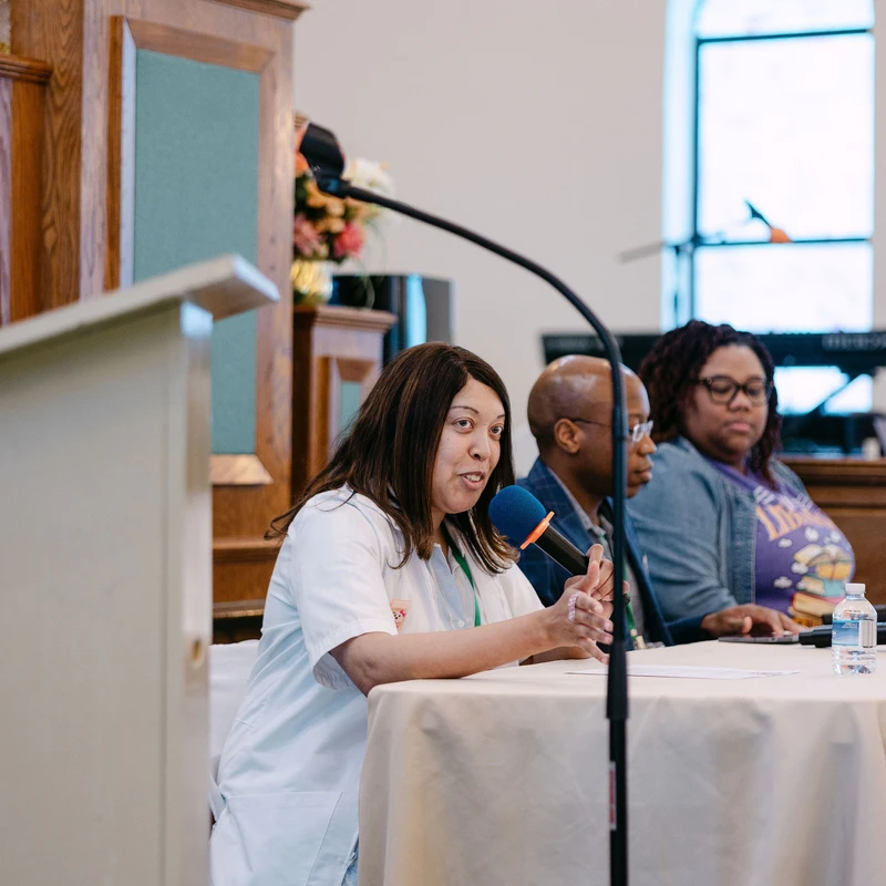 A woman speaks into a microphone at a table during a panel discussion. Two other panelists sit beside her in what appears to be a church or formal hall.