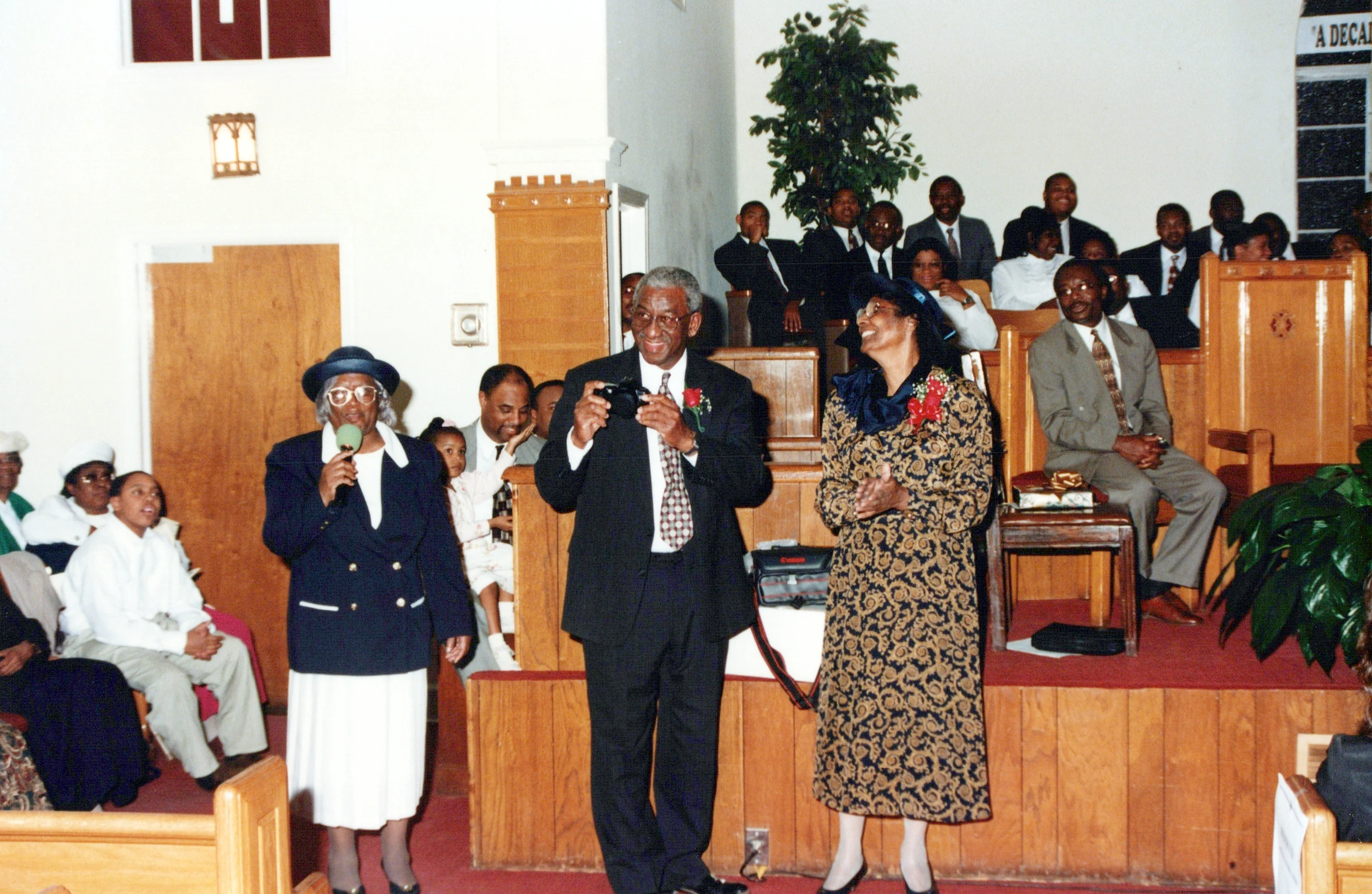 A vintage photograph of a Black congregation in a church. At the front, a woman holds a microphone, a man takes a picture, and another woman claps joyfully.