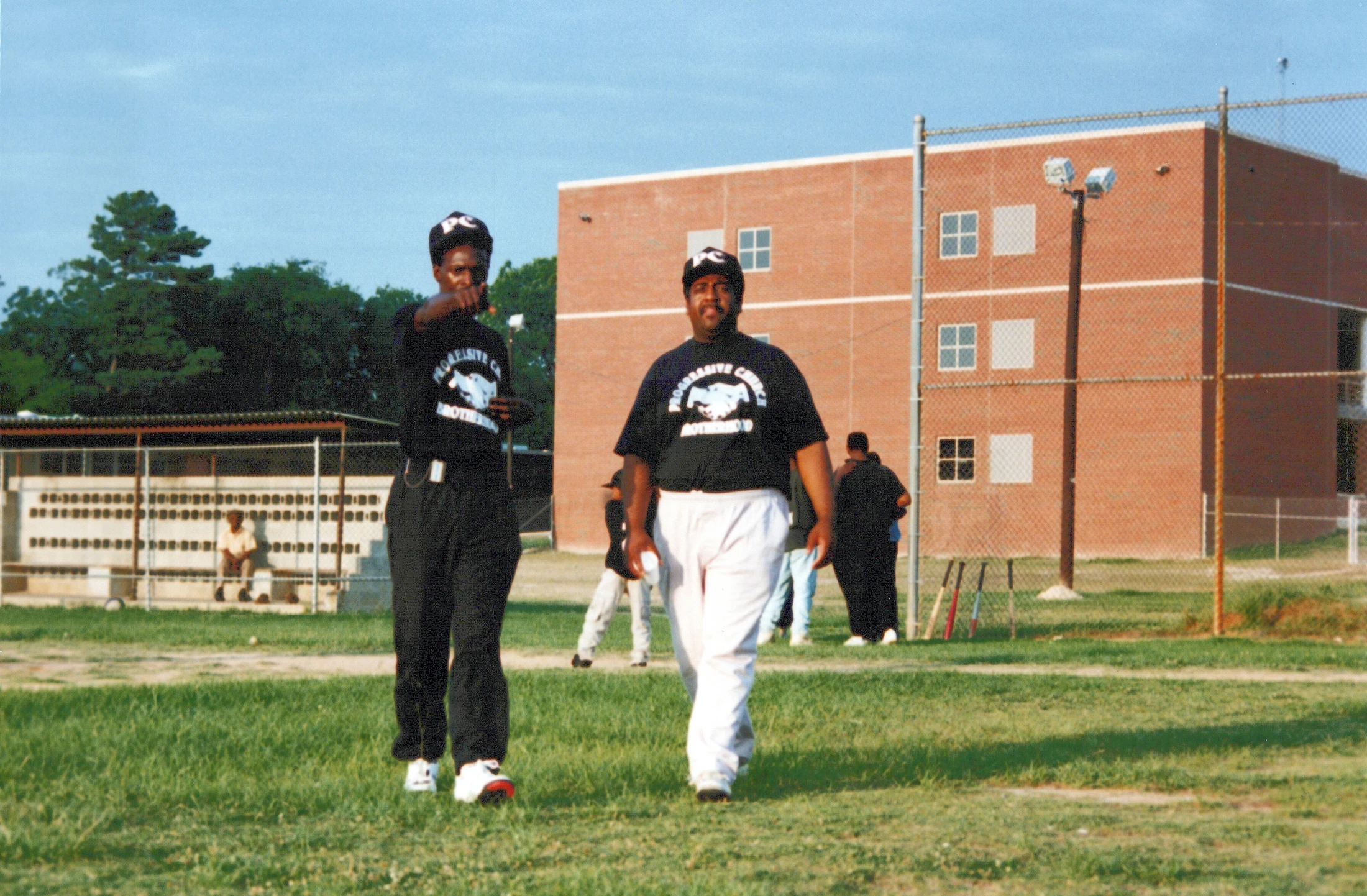 Two Black men in matching black team t-shirts walk across a grassy baseball field, with one pointing at the camera. A large brick building is in the background.