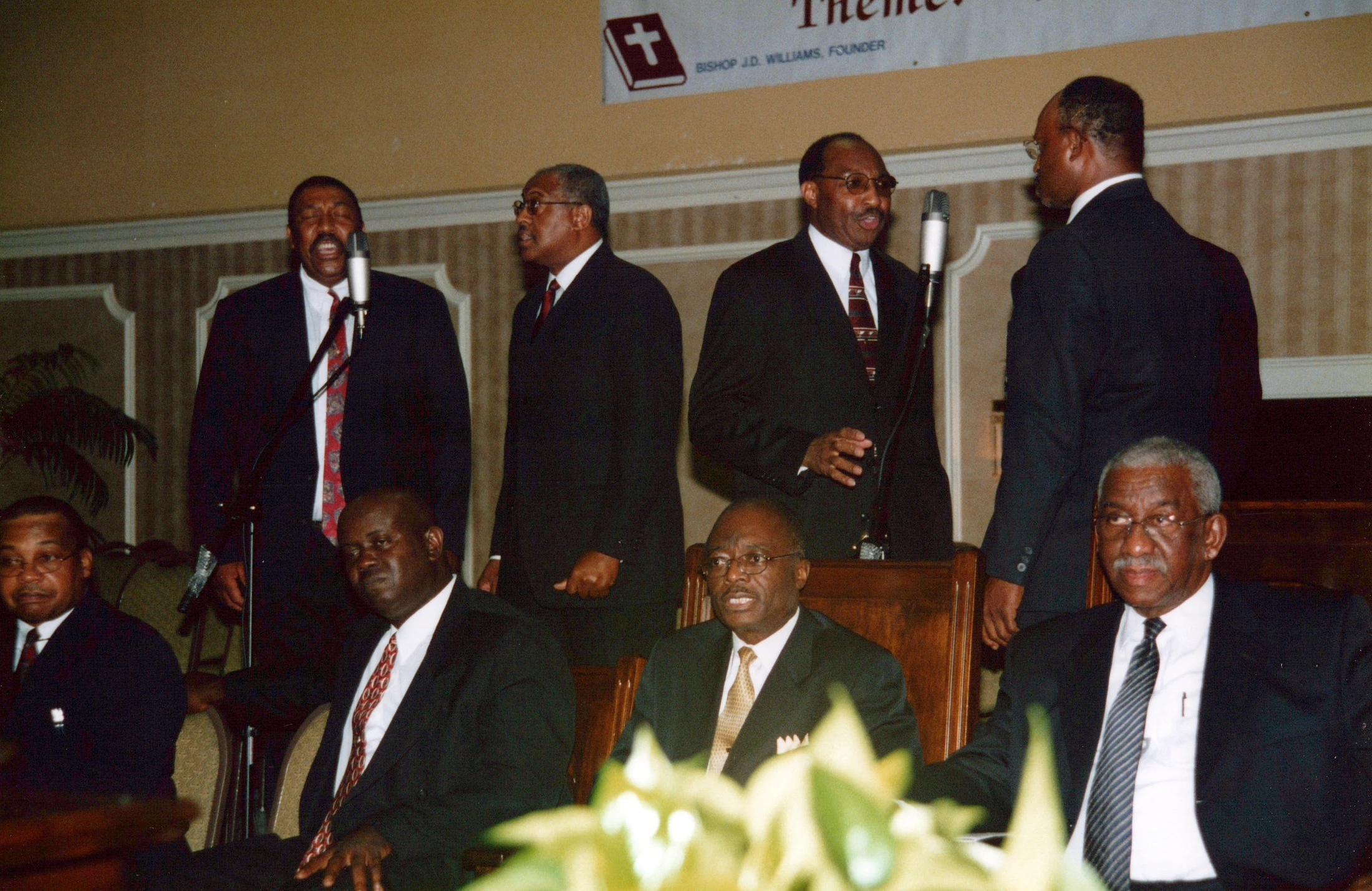 A group of eight African American men in dark suits at a church service. Several men are standing and speaking or singing into microphones.