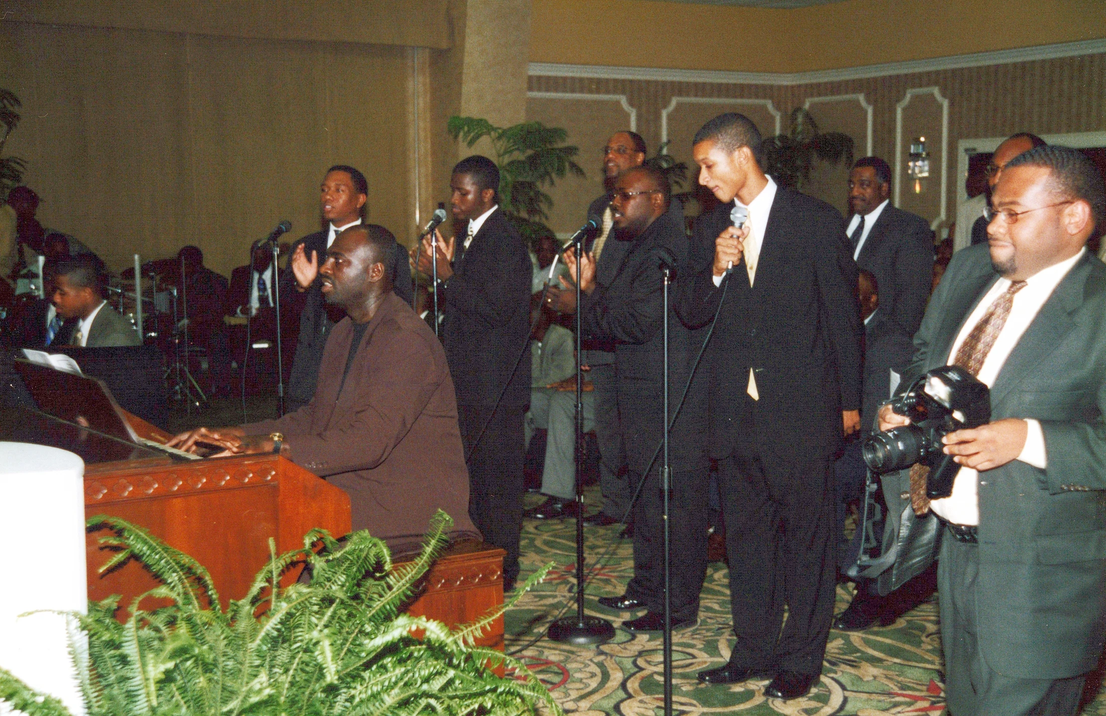 A group of Black men in suits sing into microphones. One man plays a piano while another in the foreground holds a professional camera.