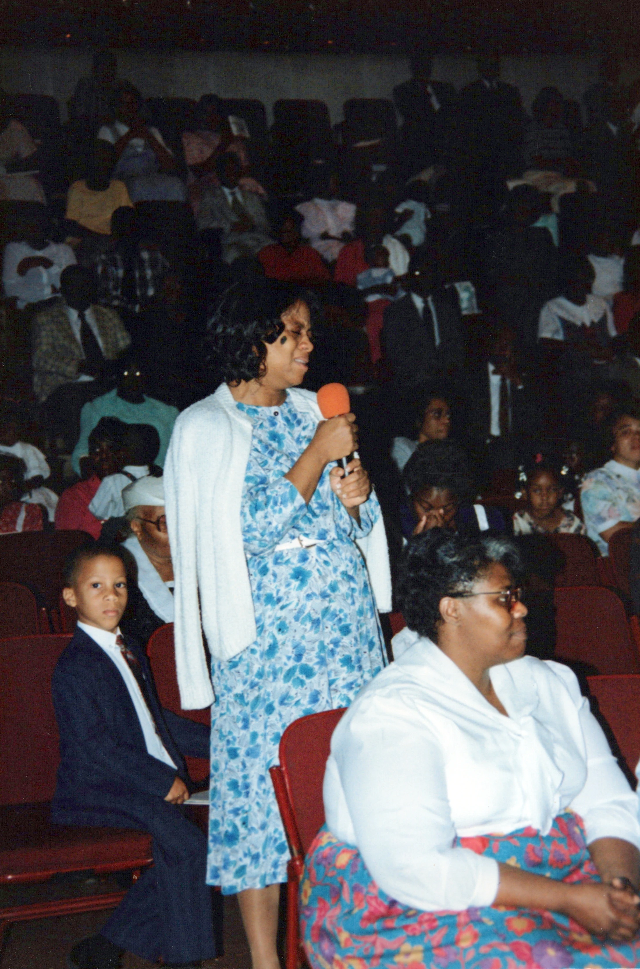 A vintage photo shows a Black woman in a floral dress speaking into a microphone while standing in an audience. A young boy in a suit sits next to her.