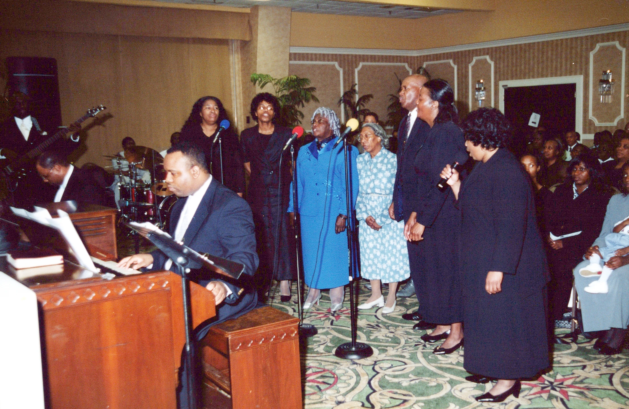 A Black gospel choir sings at microphones, accompanied by a man playing an organ, with a drummer and guitarist in a large, decorated event hall.
