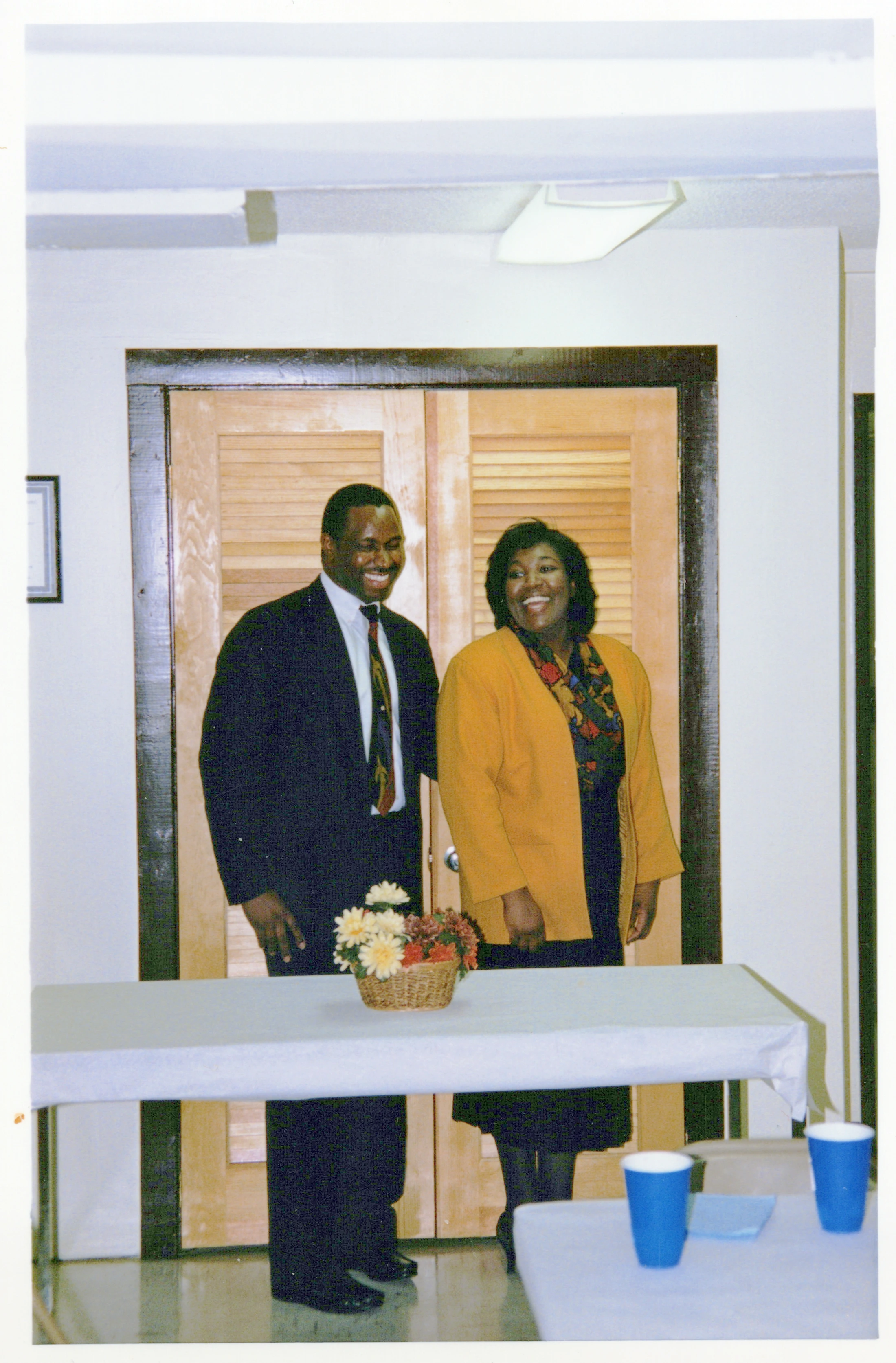 A Black man in a suit and a Black woman in a yellow cardigan stand together smiling behind a table with a flower basket at an indoor gathering.