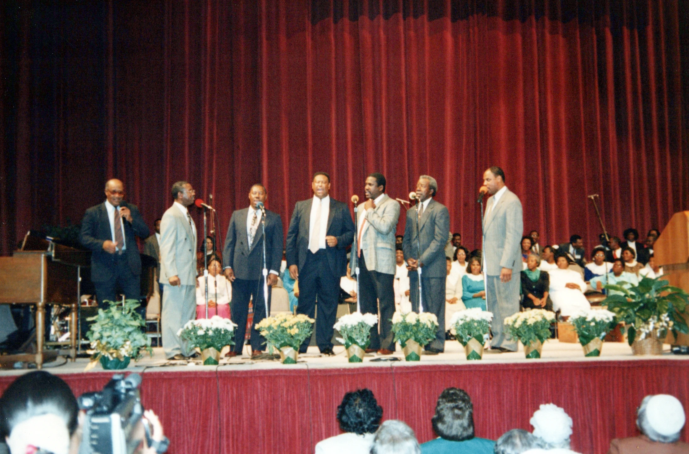 A group of seven African American men in suits sing into microphones on a stage with a red curtain, in front of an audience and potted flowers.
