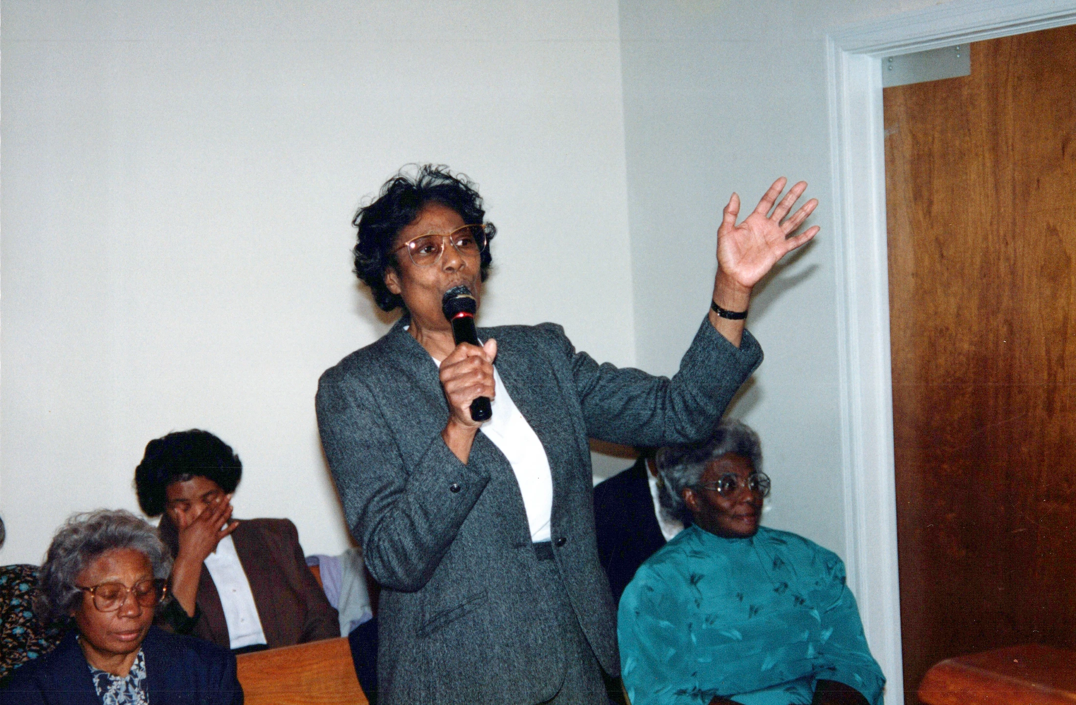 An older Black woman in a grey suit and glasses speaks into a microphone, gesturing with one hand. An audience of older women sits listening in the background.