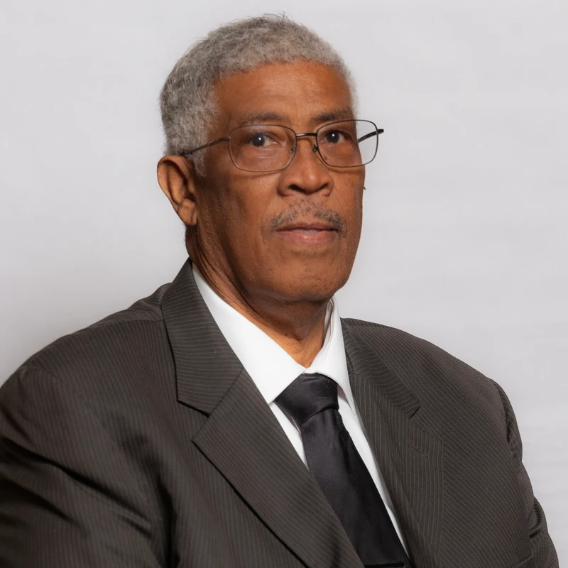 A headshot of an older African American man with gray hair and glasses. He wears a dark pinstripe suit, white shirt, and black tie against a gray background.