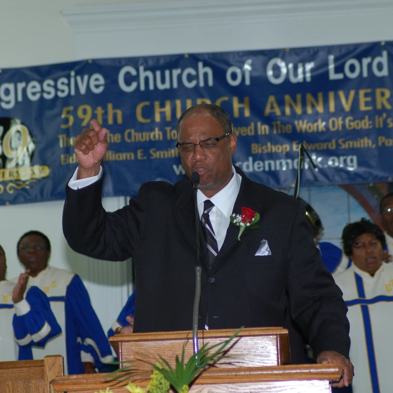 A Black pastor in a suit passionately preaches from a podium, pointing his finger up. Behind him, a choir sings in front of a 59th church anniversary banner.