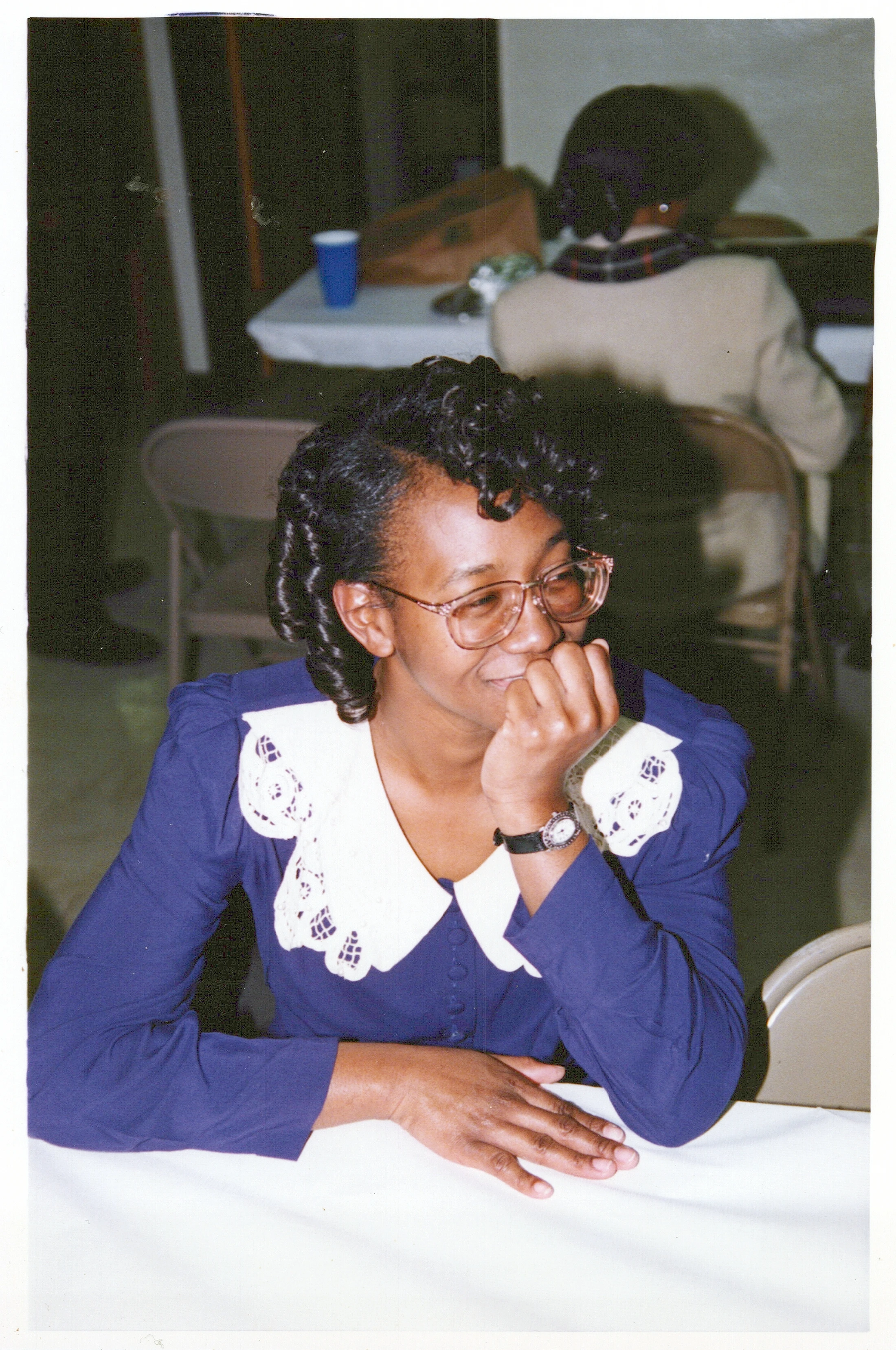 A vintage photo of a smiling Black woman sitting at a table. She wears glasses and a blue blouse with a large white lace collar, resting her chin on her hand.