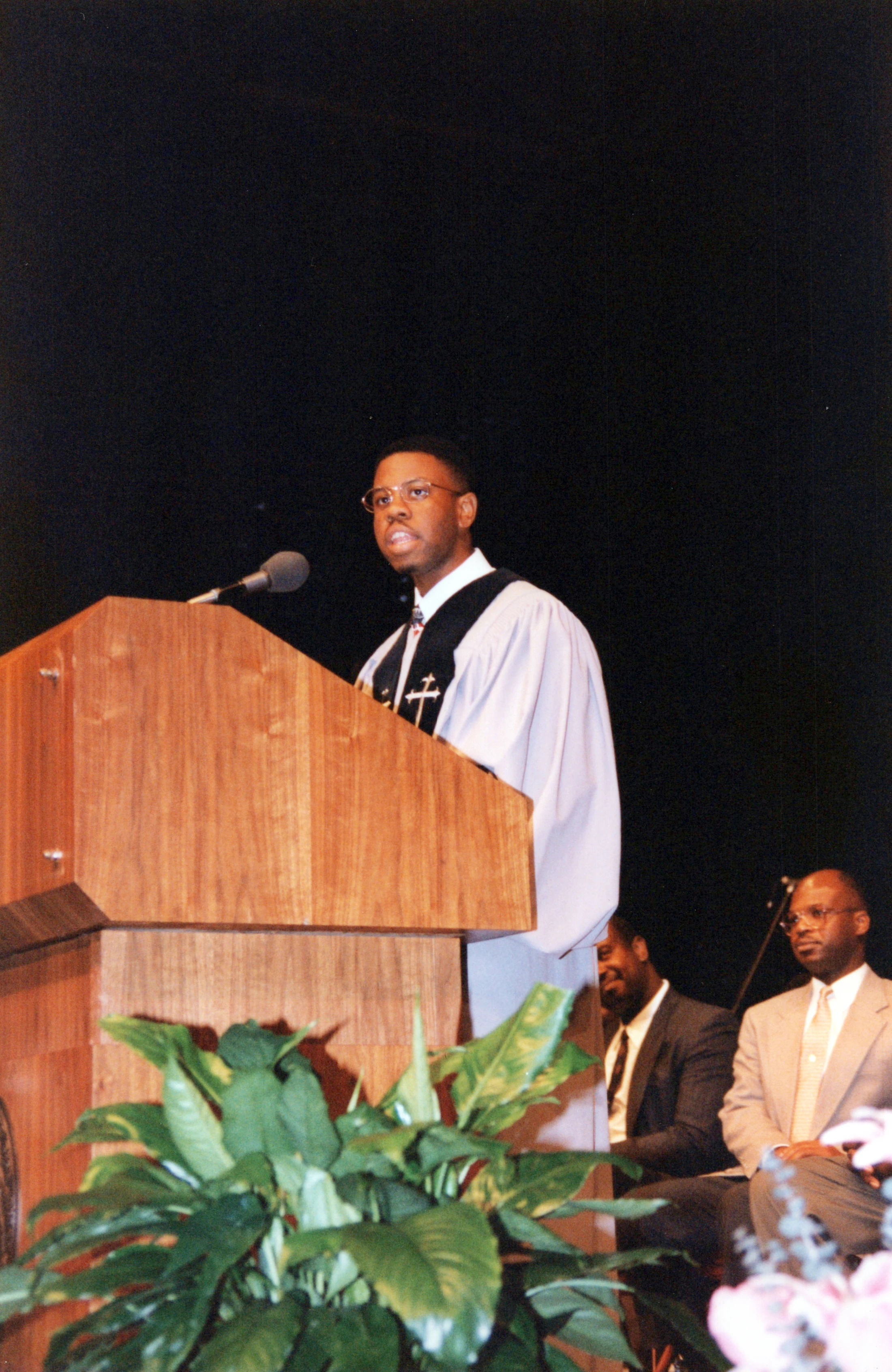 A young Black man in a preacher's robe and glasses speaks at a wooden pulpit. Two other men are seated behind him against a dark background.