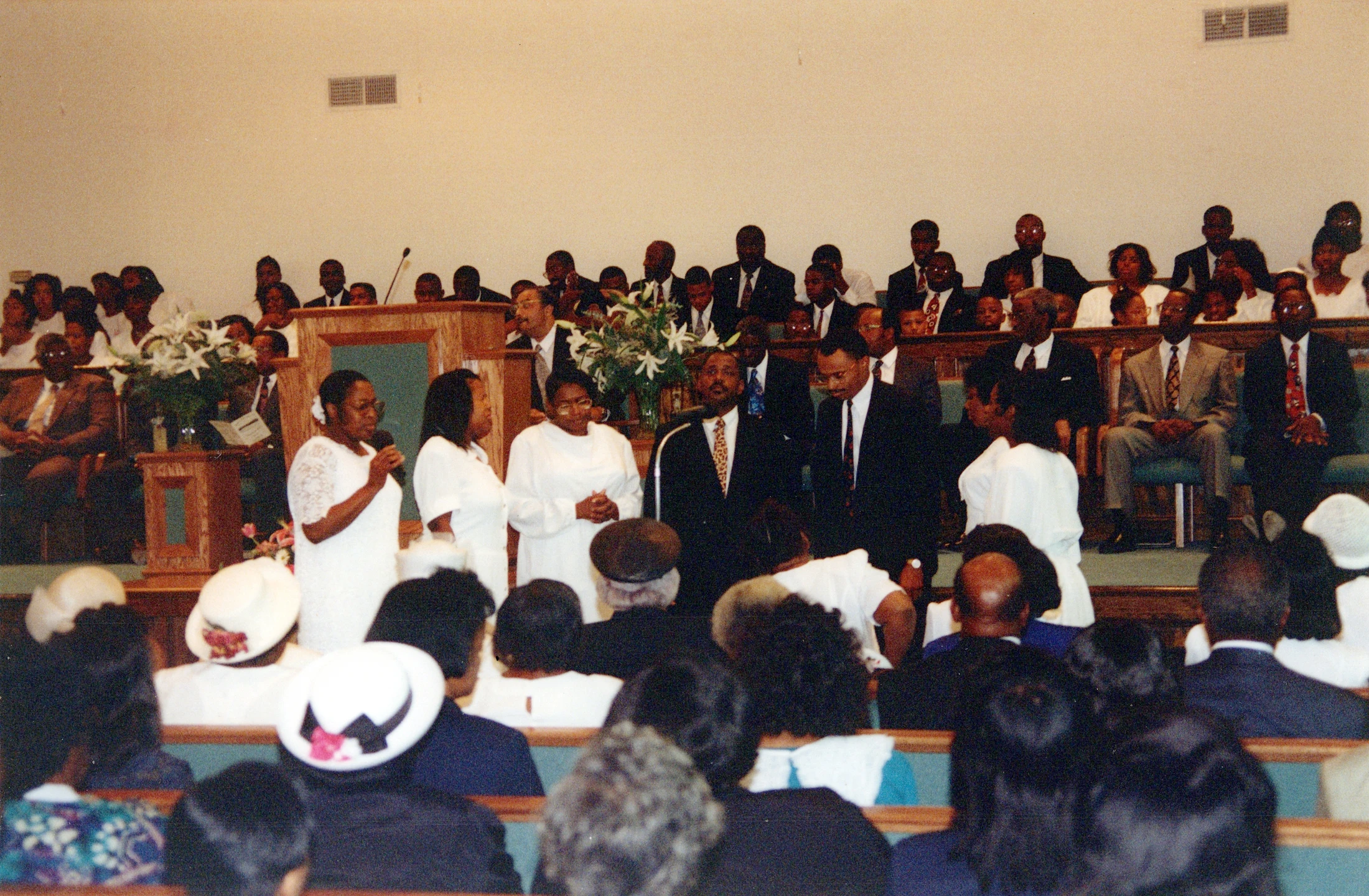A wide shot of a Black church congregation during a service. People are seated in pews, while others stand at the front in white robes and suits.