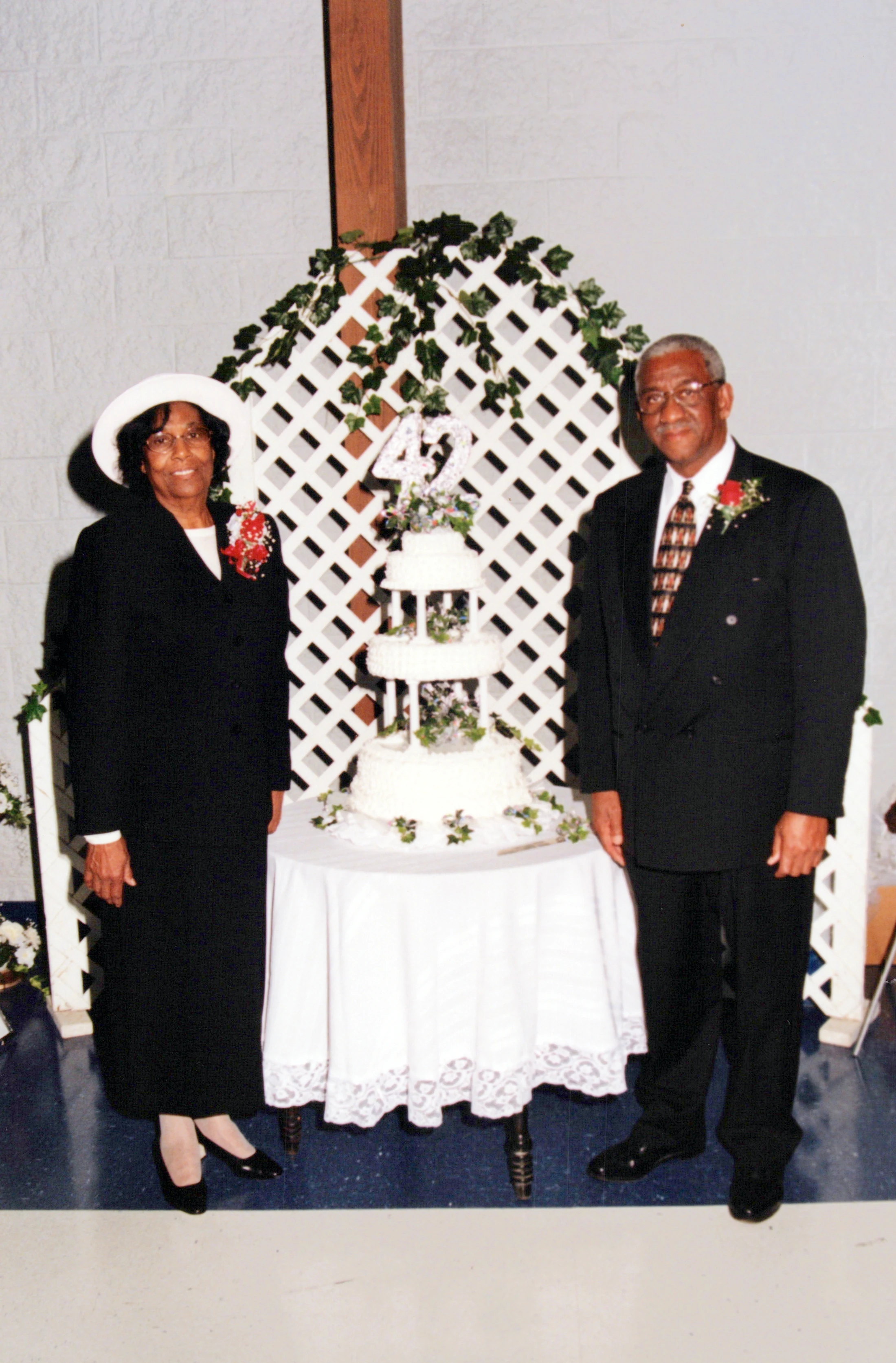An older Black couple stands on either side of a three-tiered white cake with a '42' topper, celebrating their anniversary.