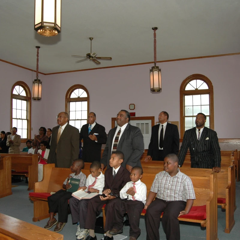 An African American congregation of men, women, and children dressed in formal attire, worshiping together in a church with wooden pews and arched windows.