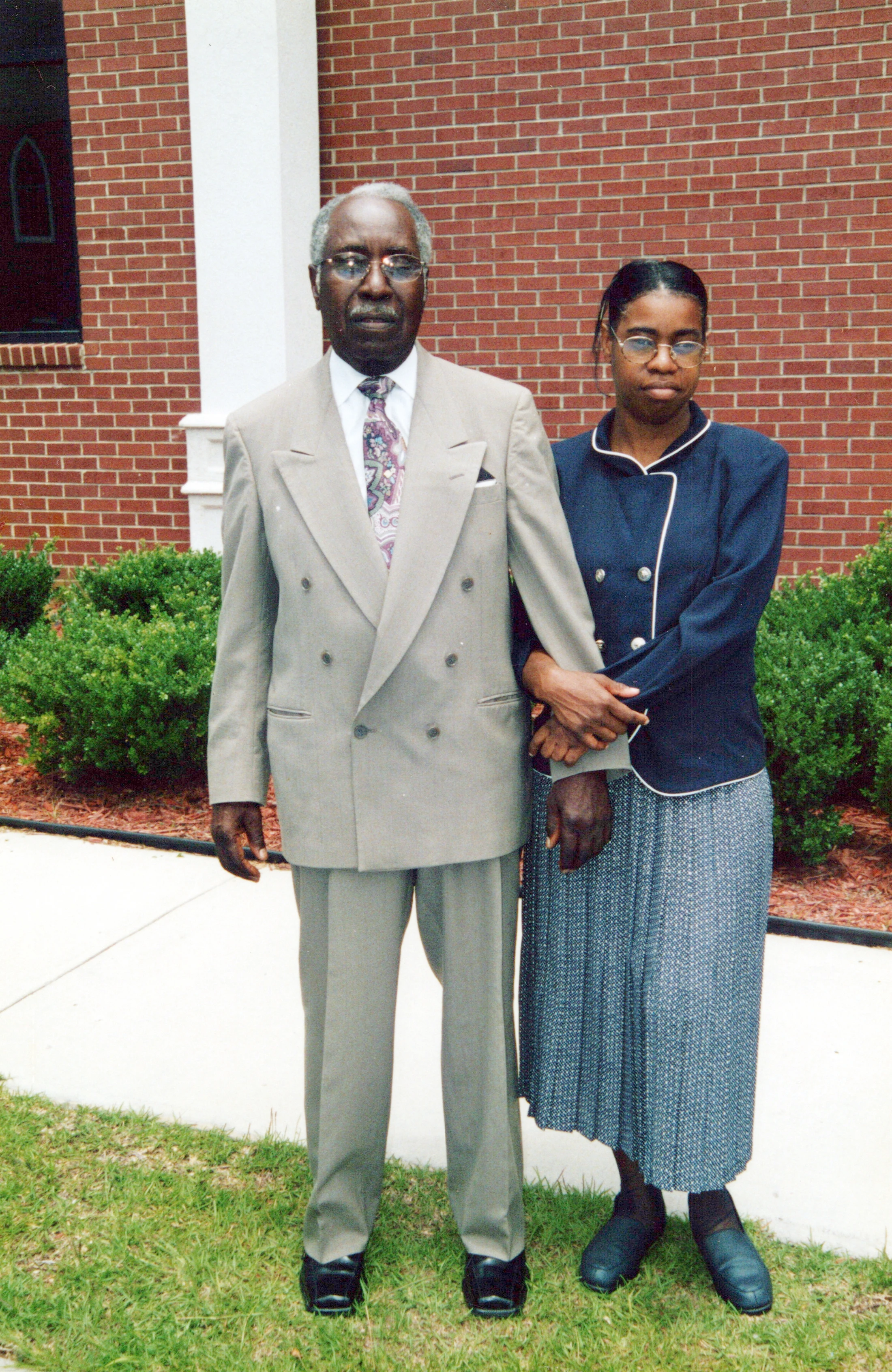 A full-length outdoor portrait of an older Black man in a suit and a younger Black woman in a jacket and skirt. They stand arm-in-arm before a brick wall.