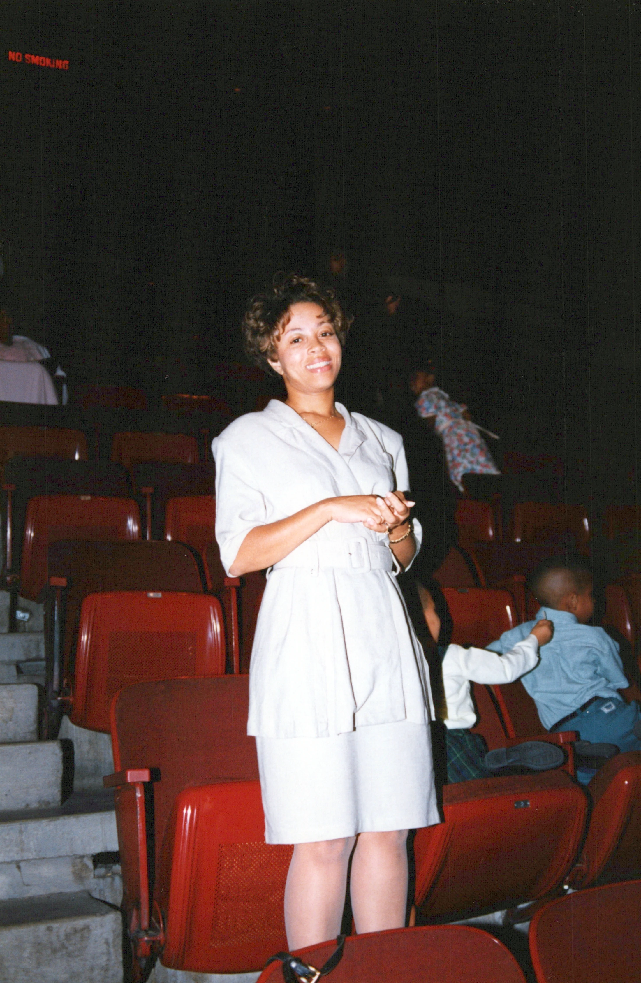 A vintage, flash photograph of a smiling woman in a white suit standing in an auditorium with rows of red seats. Other people are in the background.