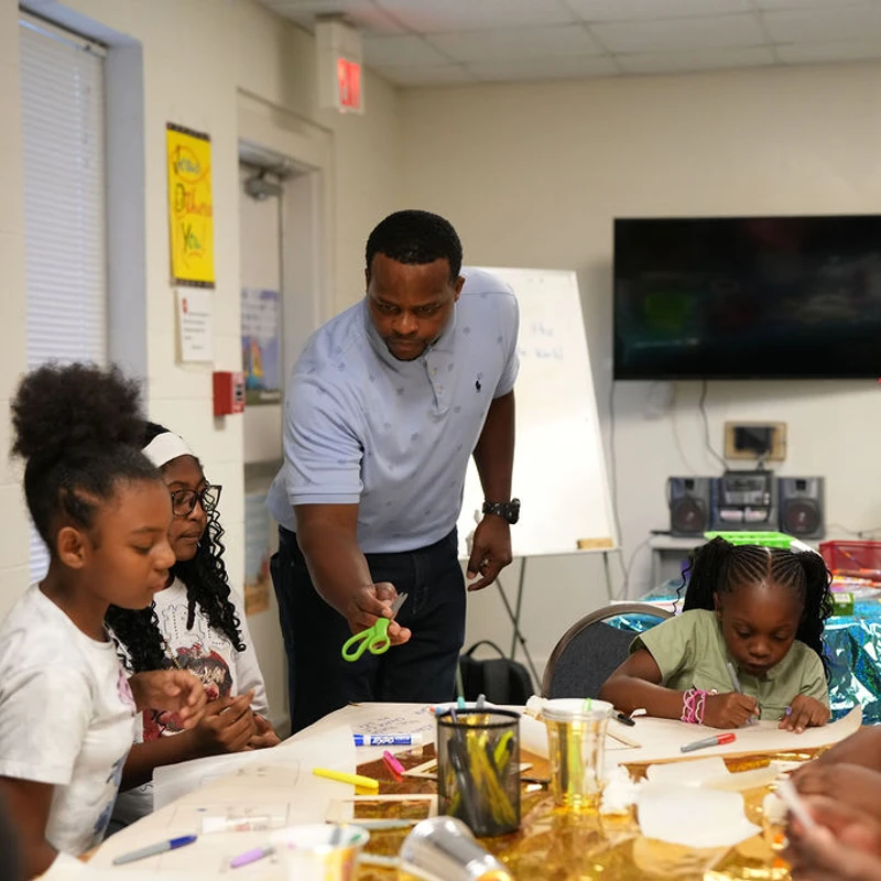 An adult male instructor leans over a table to help a group of young children with their arts and crafts projects in a classroom setting.