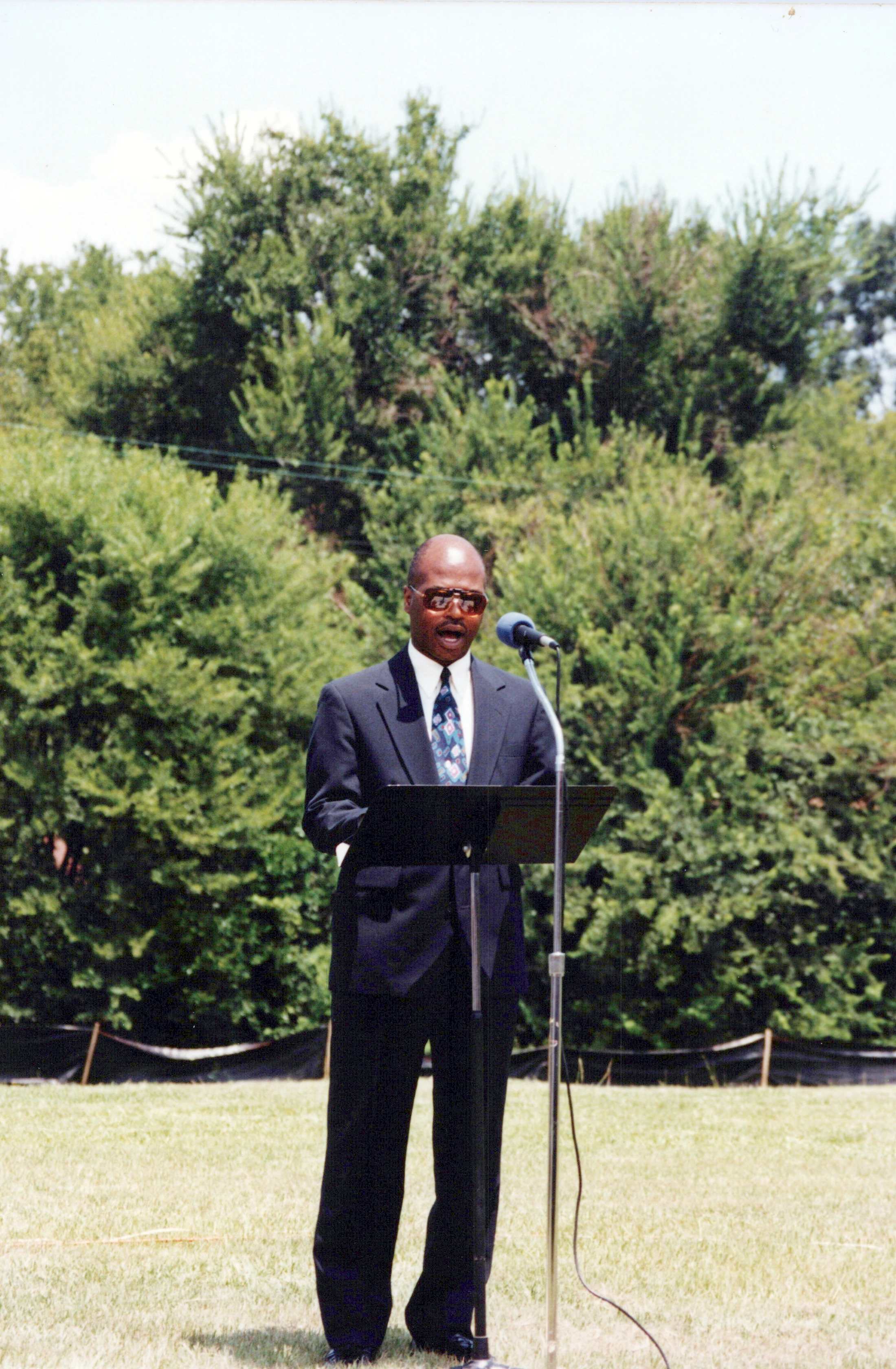 A Black man in a dark suit and sunglasses stands at a podium in a grassy field, speaking into a microphone with green trees in the background.