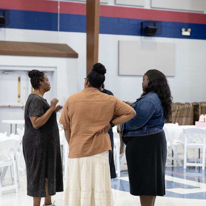 Three Black women stand in a circle talking in a large event hall with white tables and chairs. A fourth woman stands nearby.