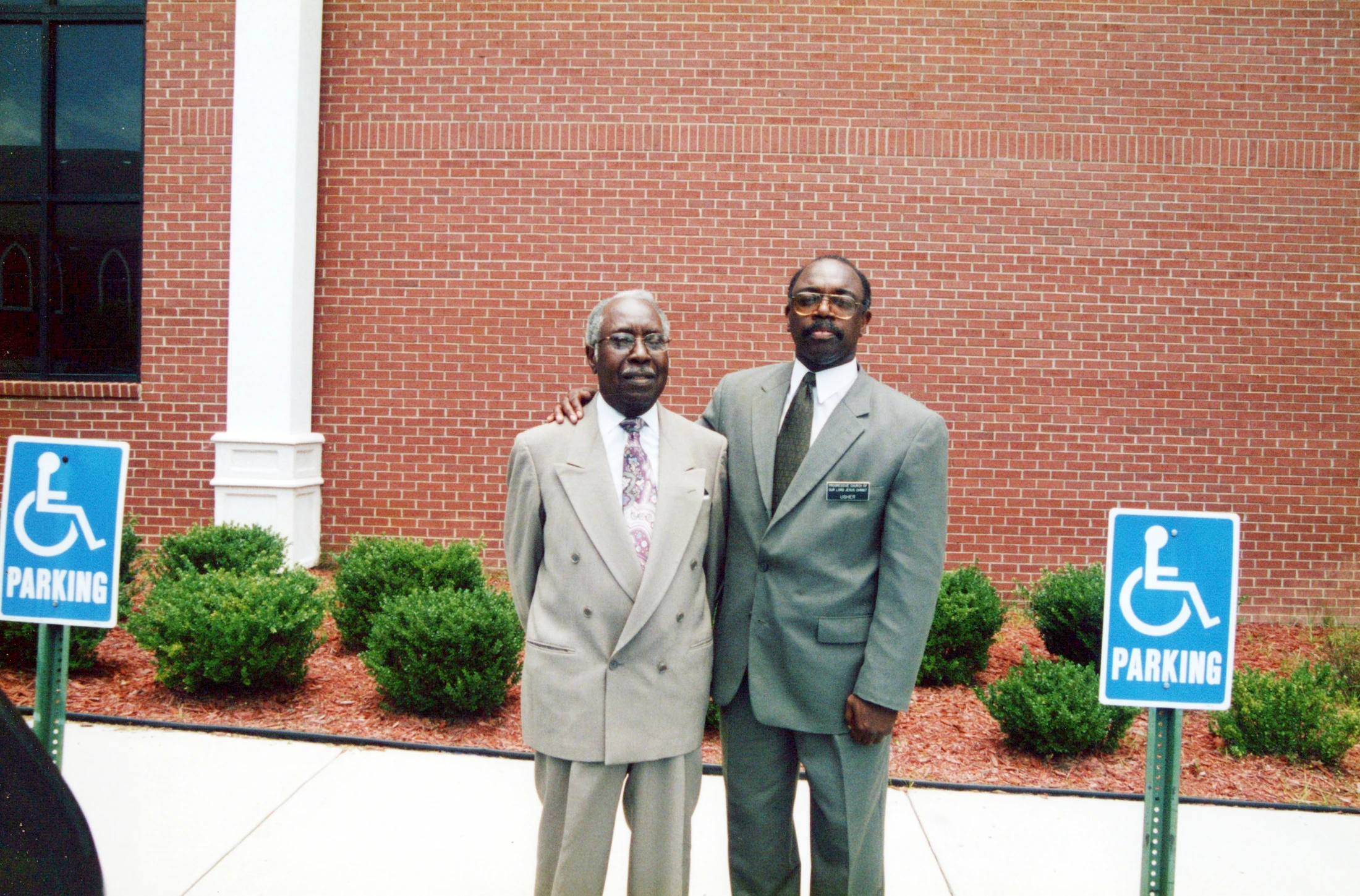 Two Black men dressed in suits stand side-by-side for a photo in front of a red brick wall, with handicapped parking signs on either side of them.