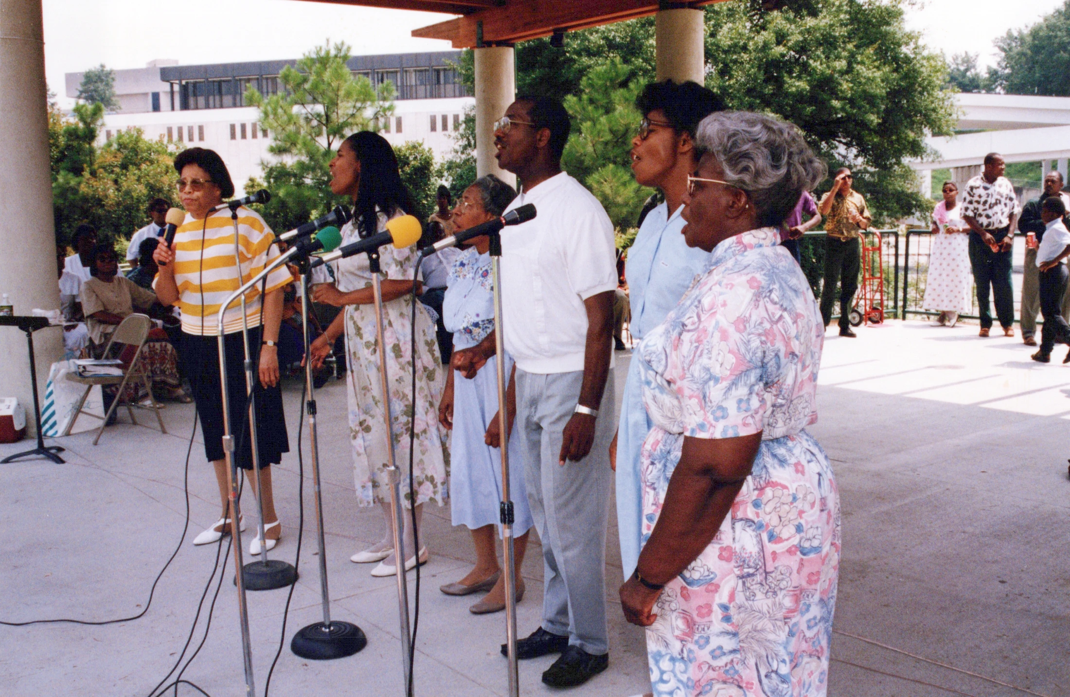 A group of African American men and women sing into microphones while performing outdoors as a choir on a concrete patio.