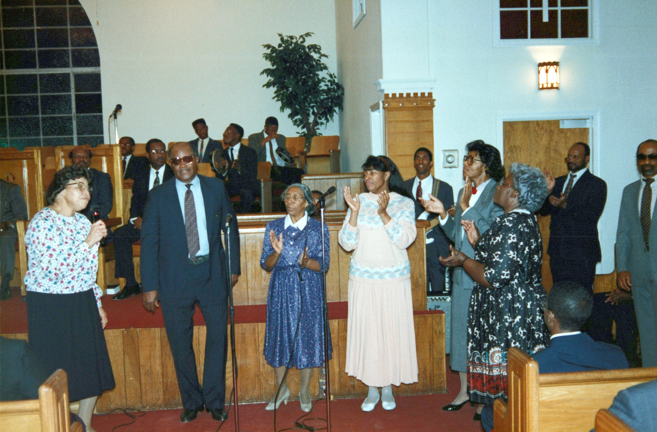 A group of African American men and women stand on a wooden platform in a church, dressed in suits and dresses, clapping their hands in joyful worship.