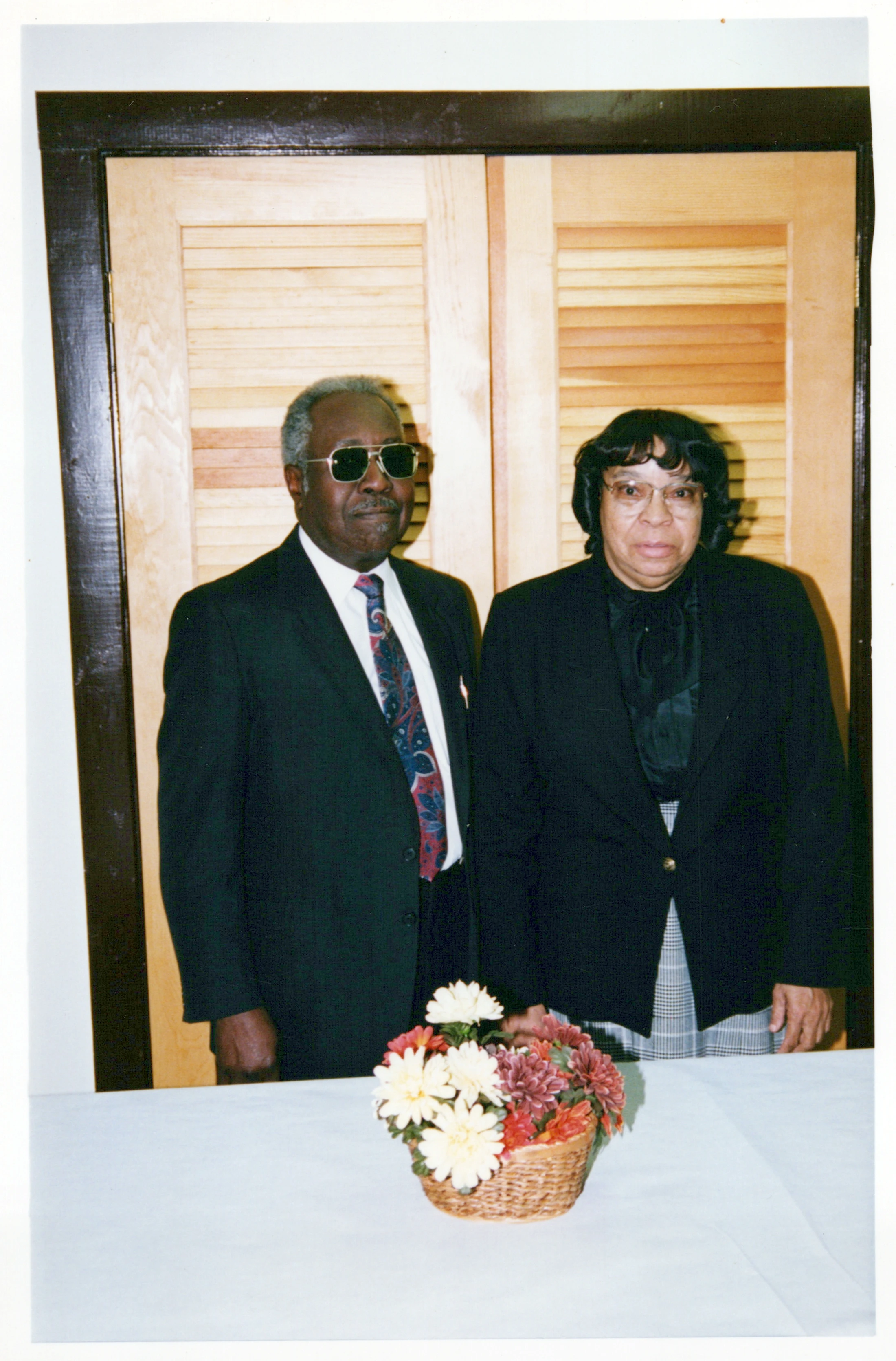 An older Black man in a suit and sunglasses stands next to a woman in a blazer. They are behind a table with a basket of flowers.