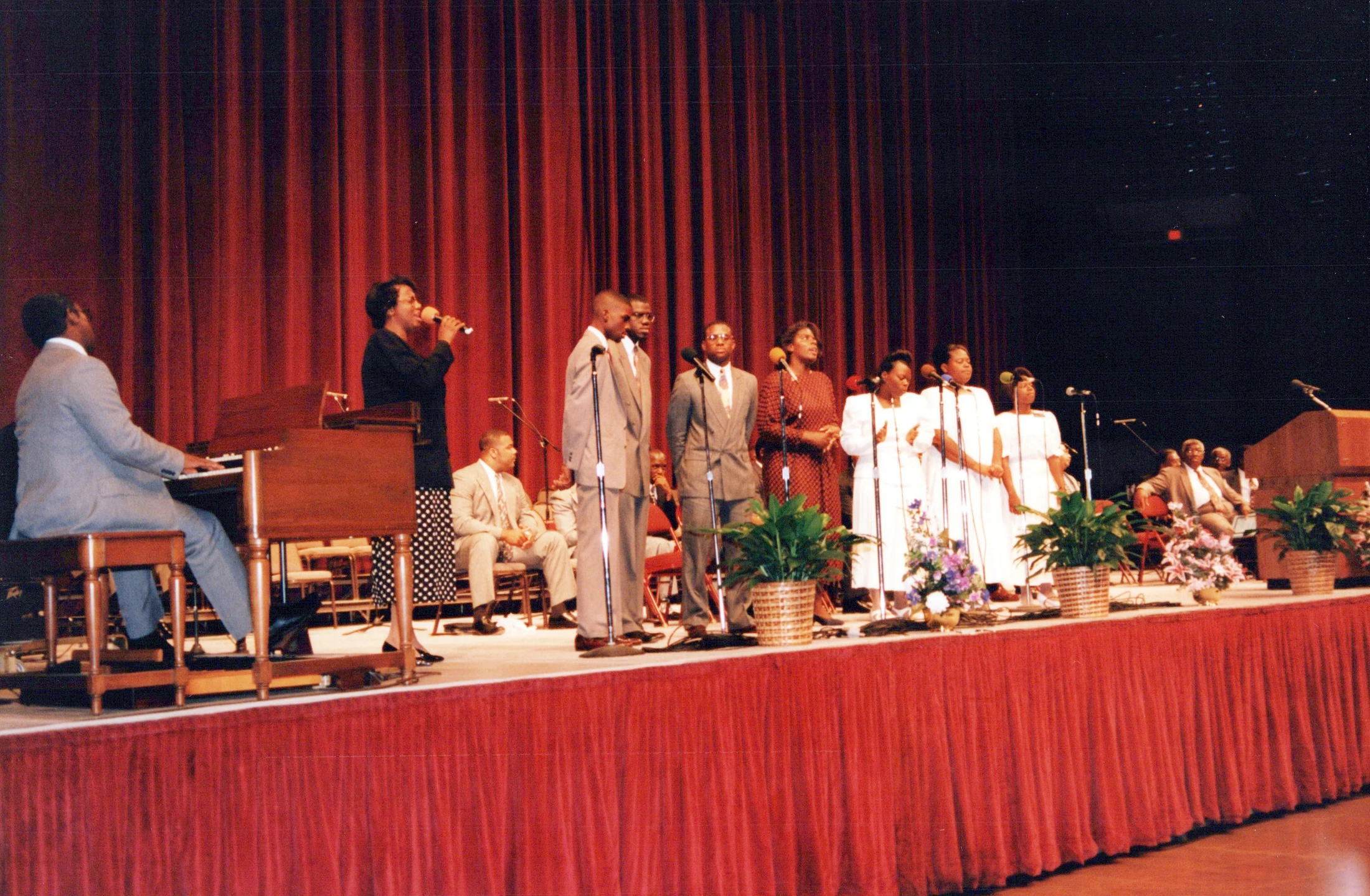 An African American gospel choir performs on a stage with a red curtain. A man plays an organ as singers in suits and white robes sing into microphones.
