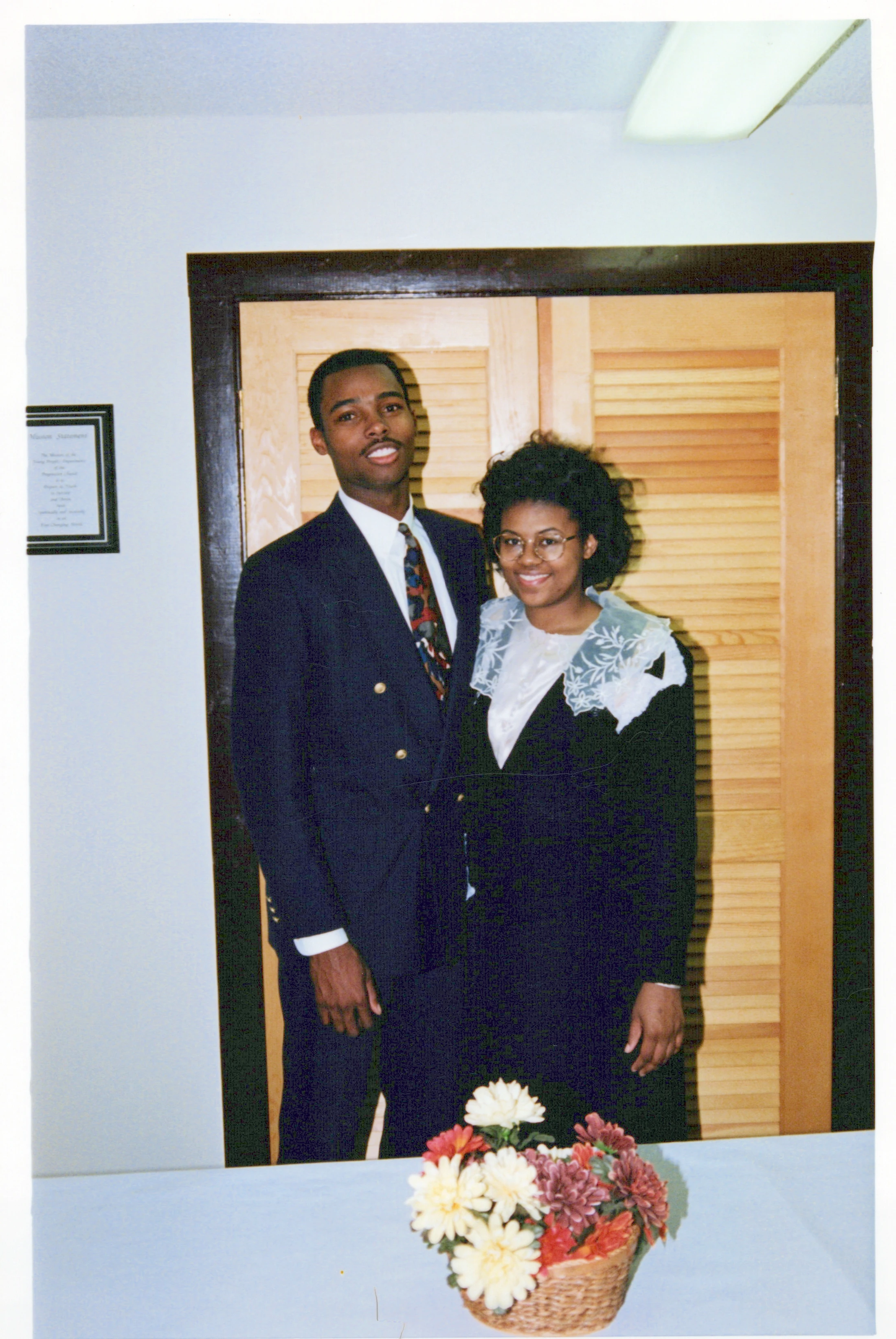 A vintage photograph of a young Black man and woman dressed in formal suits, standing side-by-side and smiling for the camera in an indoor setting.