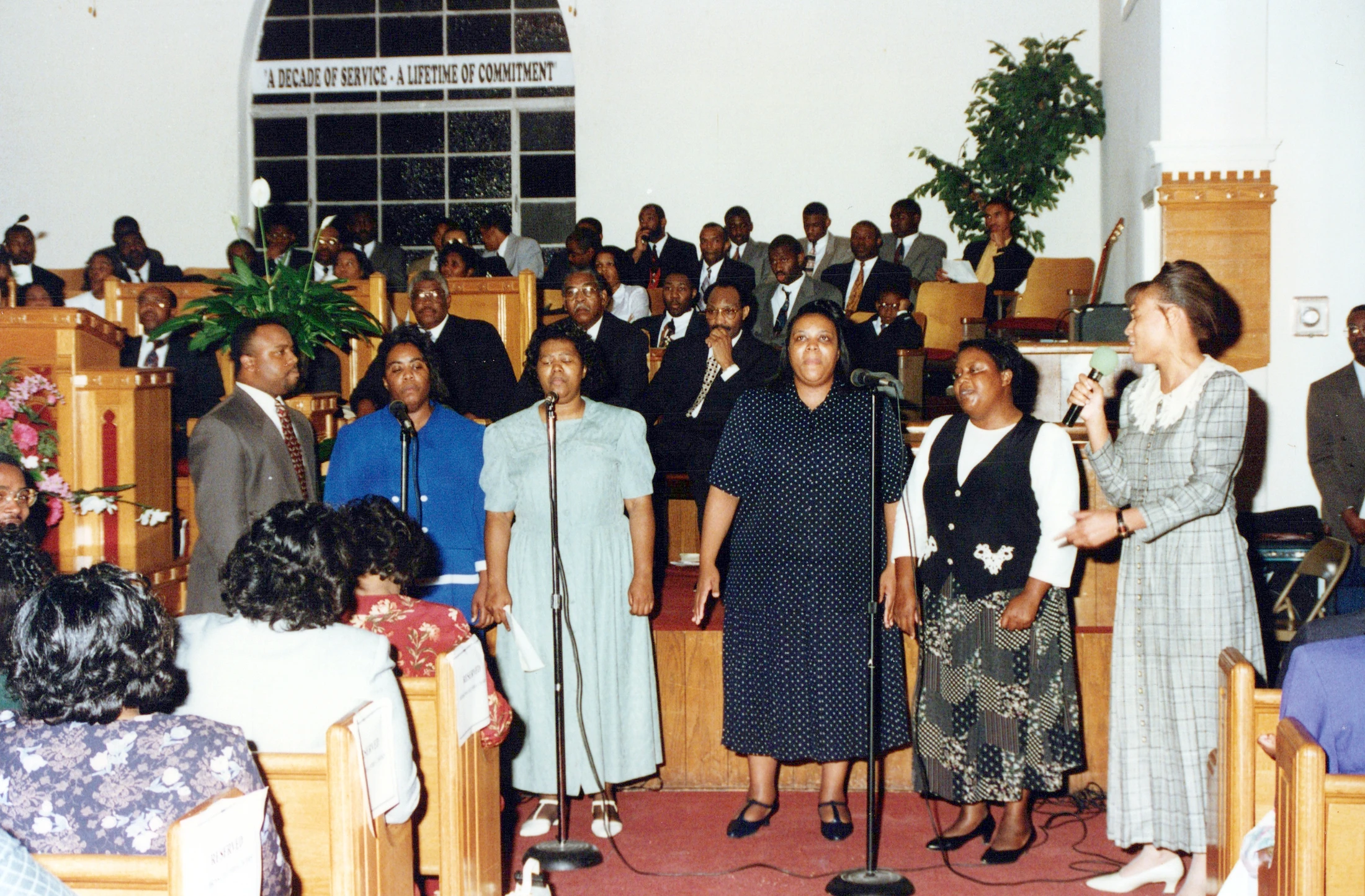 A vintage photograph shows a Black church choir of four women singing at microphones, led by another woman. The congregation is seated in pews behind them.