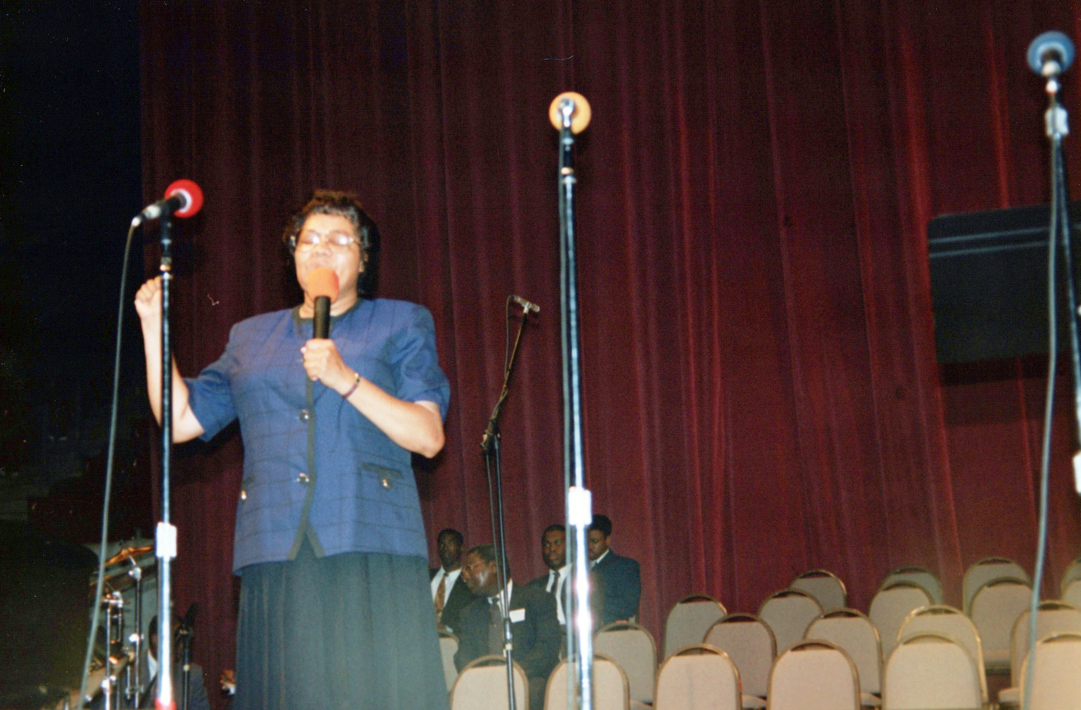 An older Black woman wearing glasses and a blue jacket speaks into a microphone on a stage. Behind her is a red curtain and several men in suits are seated.