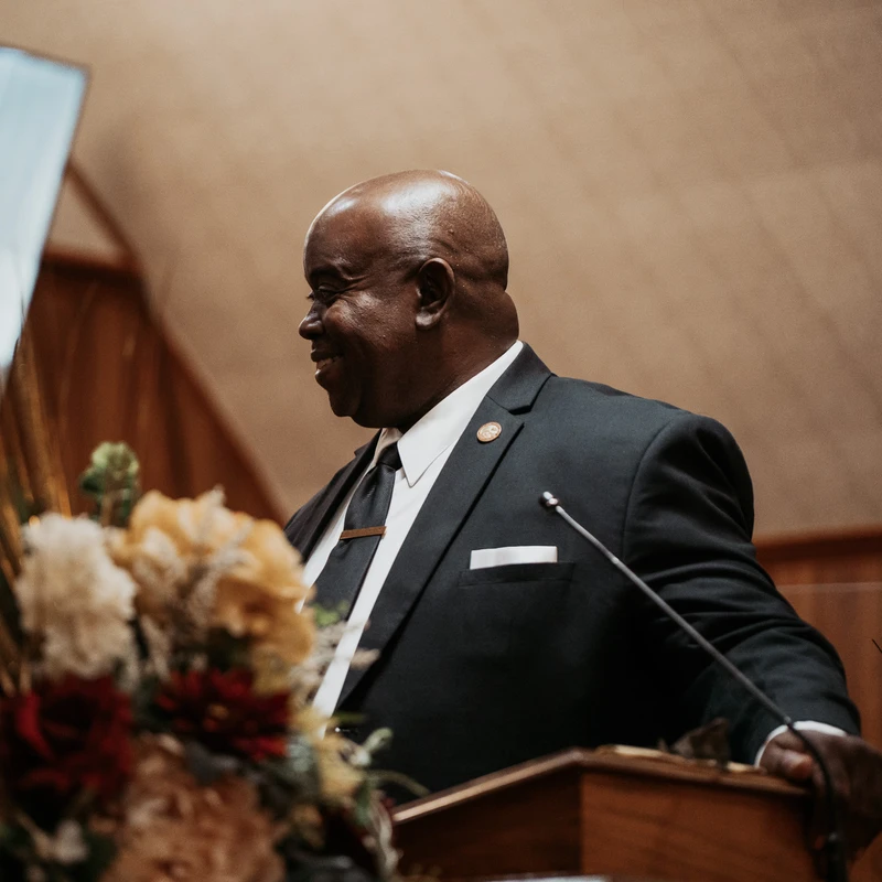A smiling, bald Black man in a dark suit and tie stands behind a wooden podium, with a microphone and blurred flowers in the foreground.