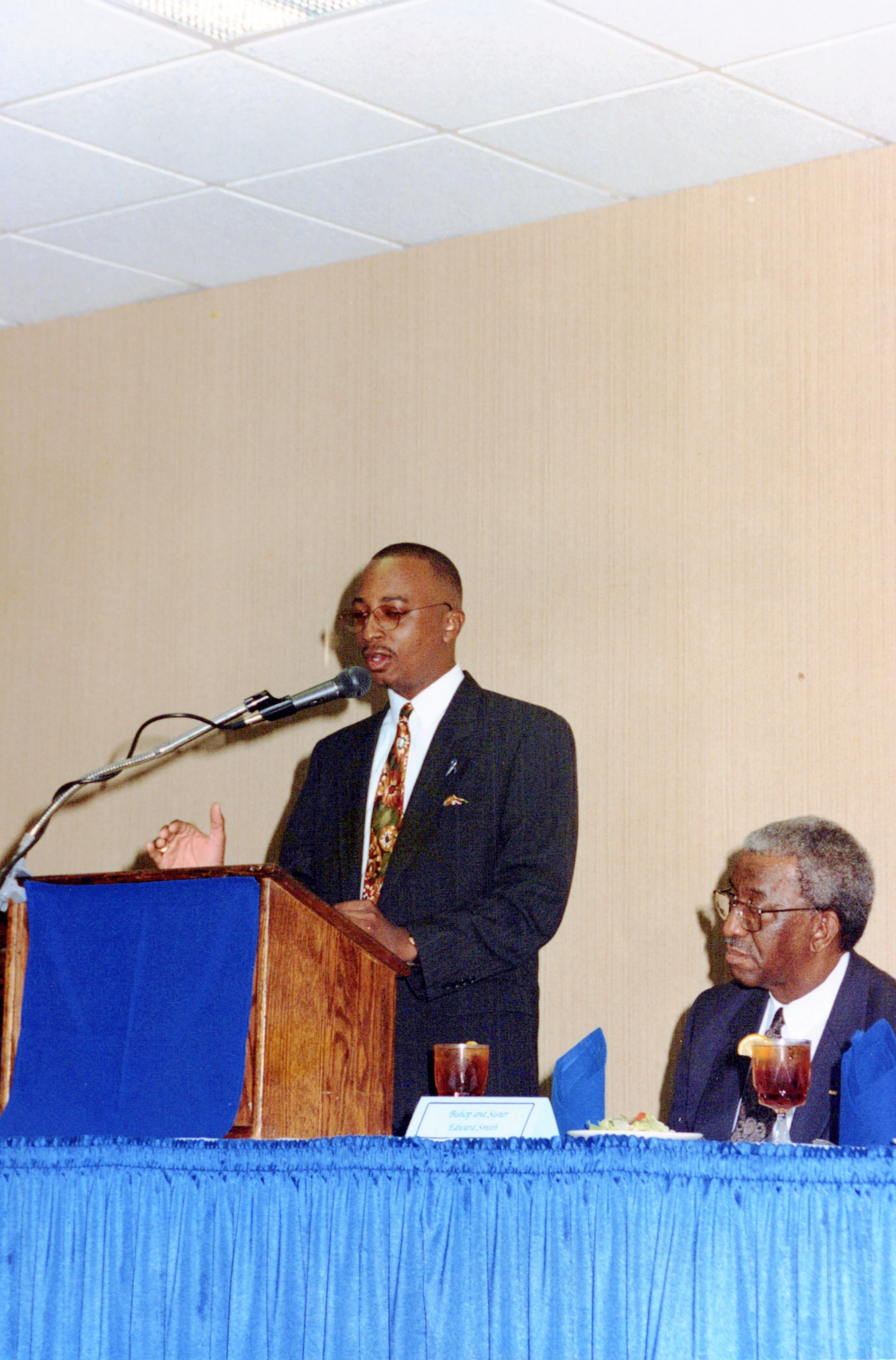 An African American man in a dark suit and glasses speaks at a wooden lectern. Another man sits at a blue-covered table beside him, listening.