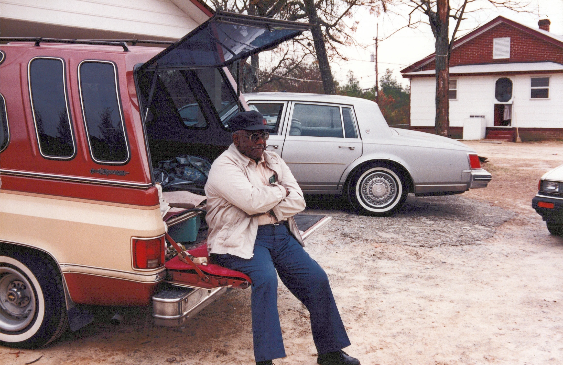 An older Black man in a cap and sunglasses sits with his arms crossed on the open tailgate of a red and tan vintage truck in a gravel driveway.