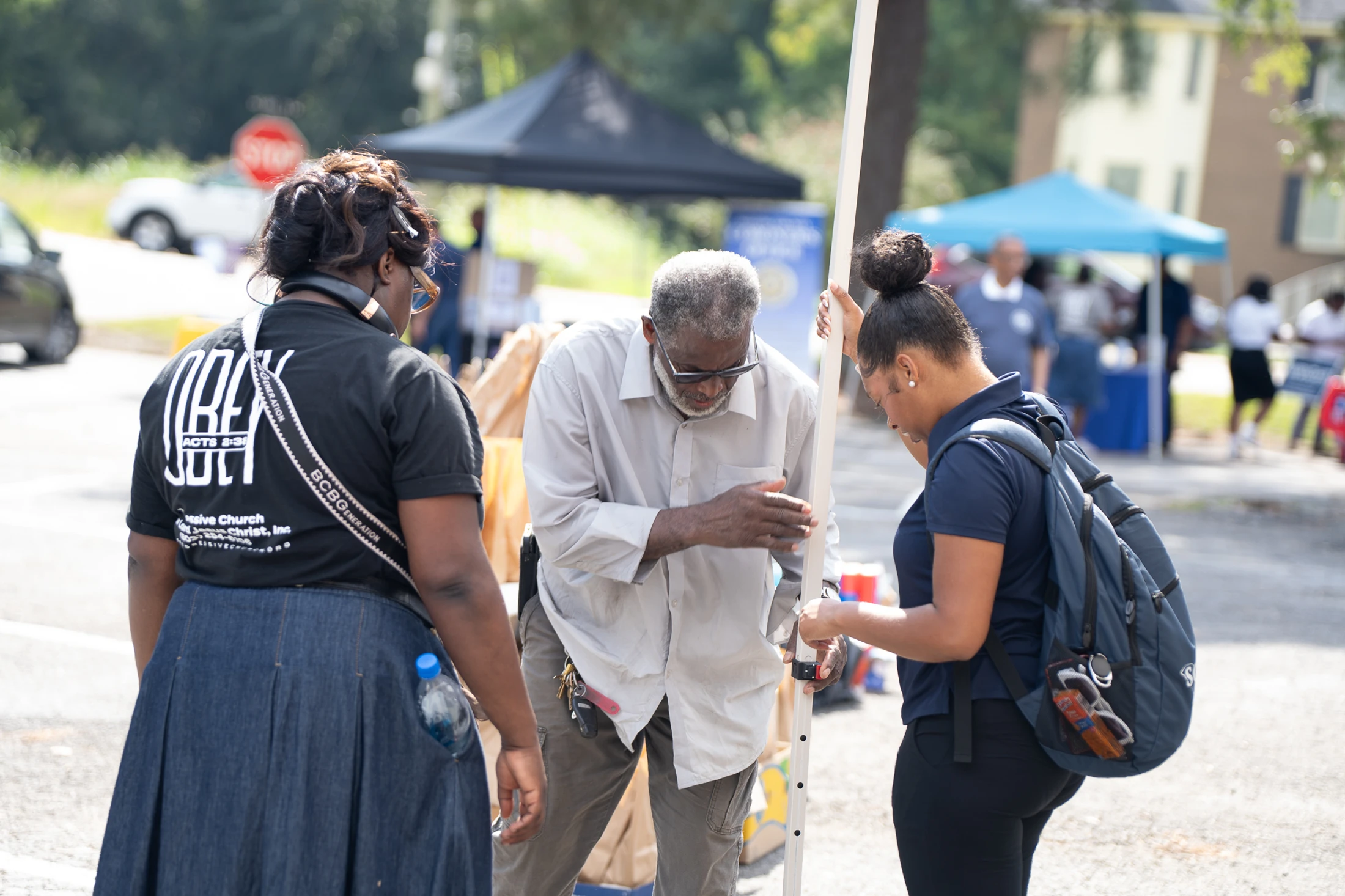 An older Black man in a light-colored shirt is assisted by two younger Black women at a sunny outdoor community event with tents in the background.