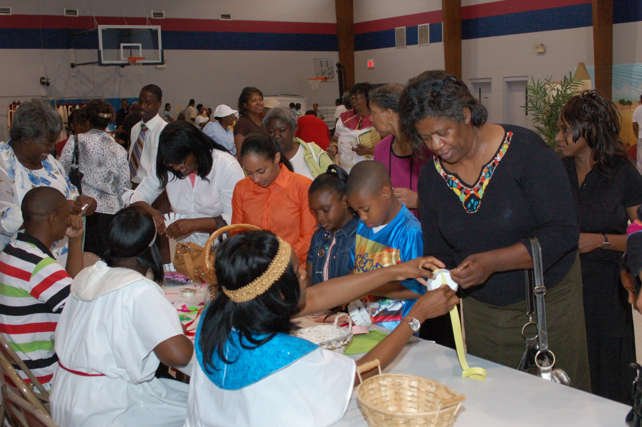 A diverse group of African American adults and children gather around a long table in a gymnasium, interacting and working on a craft project together.