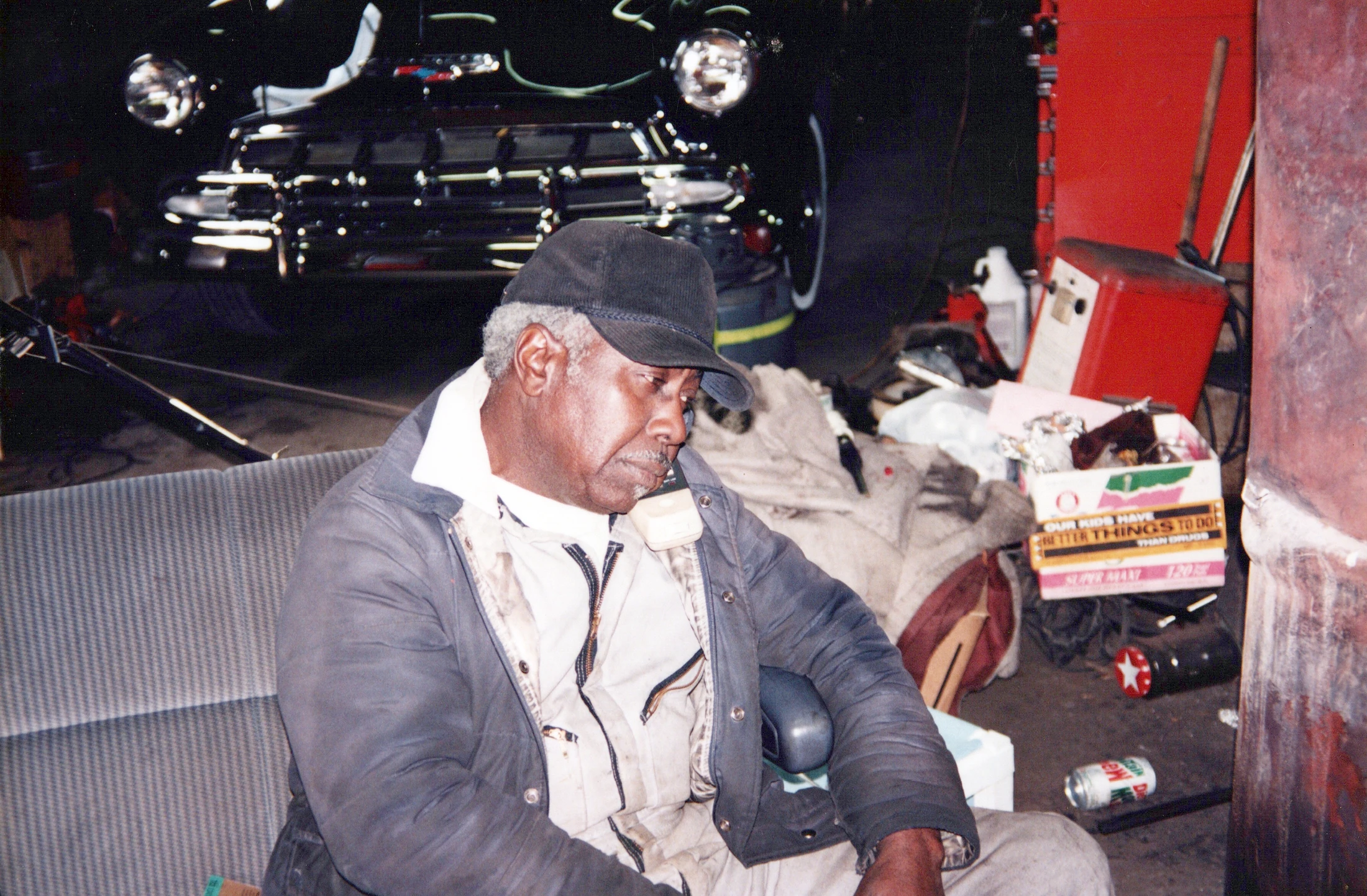 An older Black man wearing a cap and jacket sits on a car seat inside a garage, looking down thoughtfully. A classic black car is parked in the background.