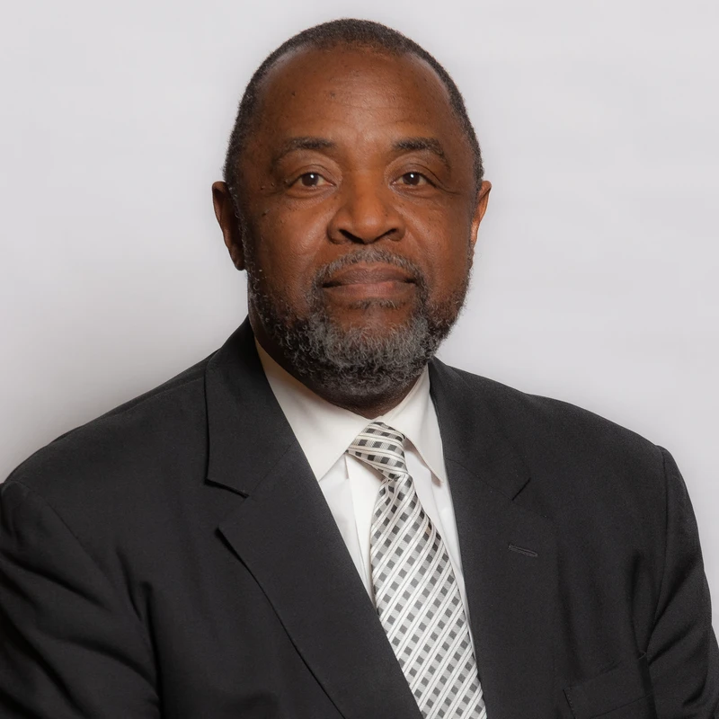 A head-and-shoulders portrait of a middle-aged Black man with a graying beard, wearing a black suit, white shirt, and patterned tie against a plain background.