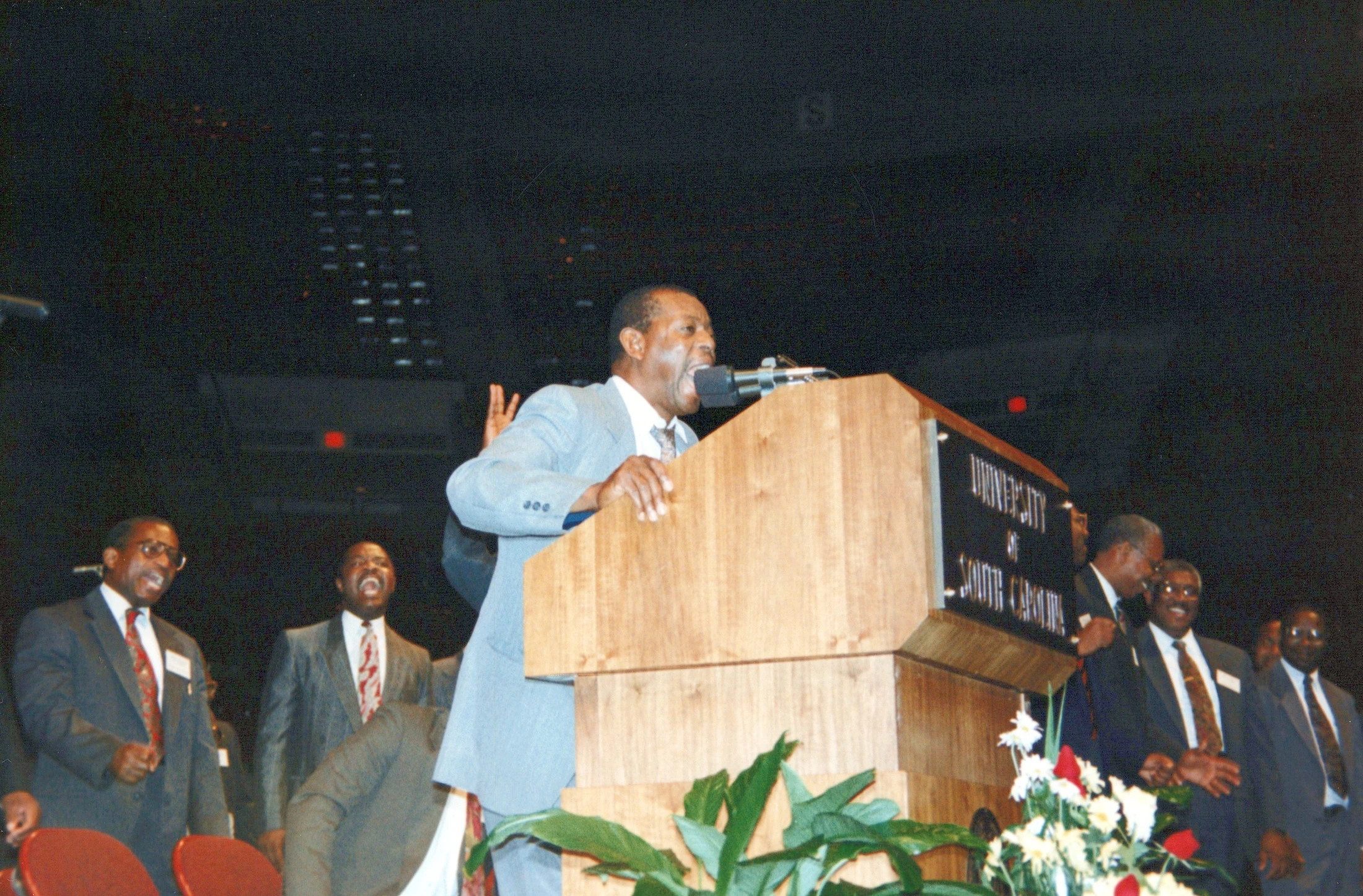 A Black man in a light-colored suit speaks passionately into a microphone at a wooden podium. Other men in suits watch and react in the background.