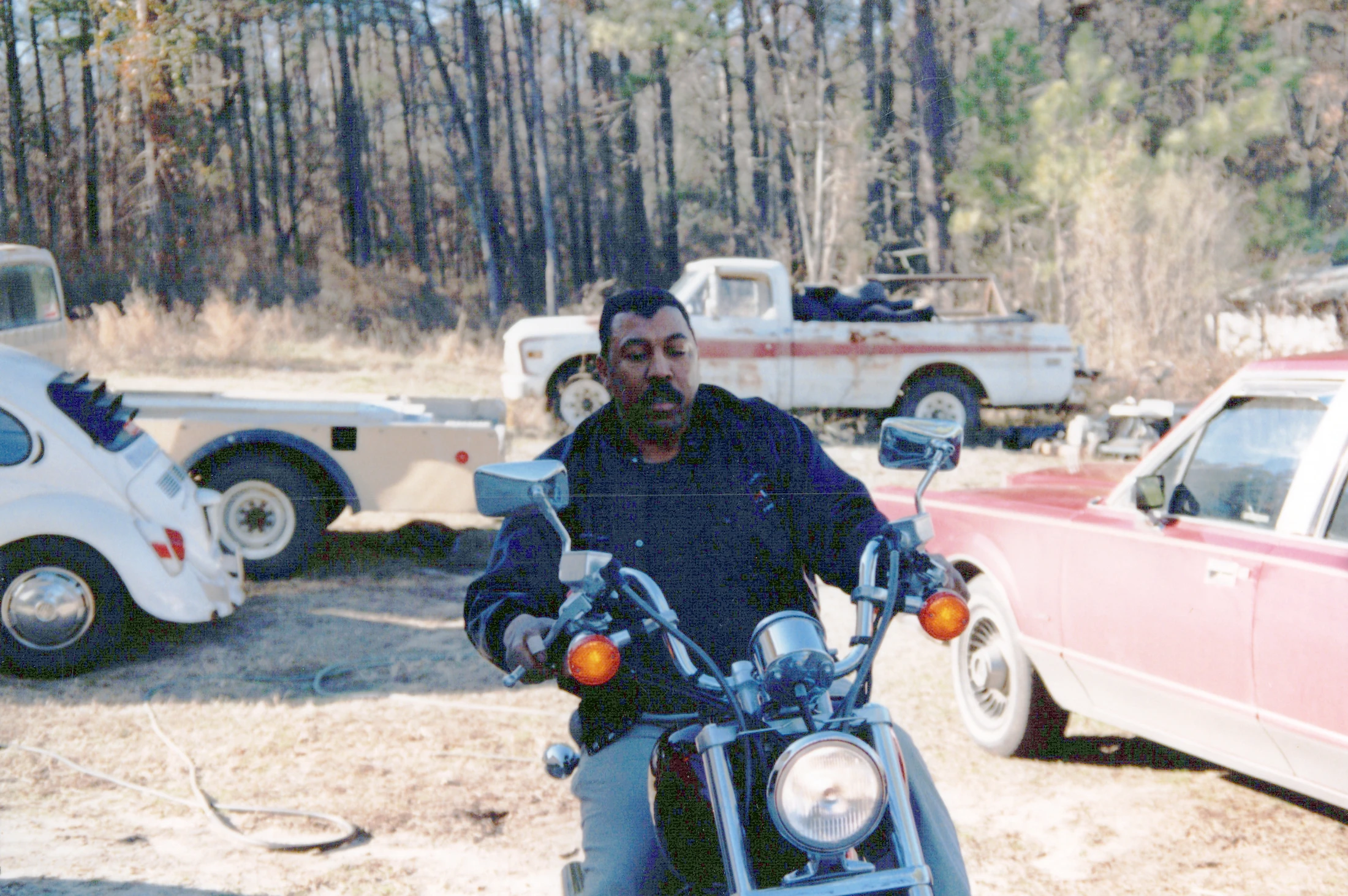 A man with a mustache sits on a motorcycle in a grassy area. In the background are several other cars, including a pickup truck, and a line of trees.
