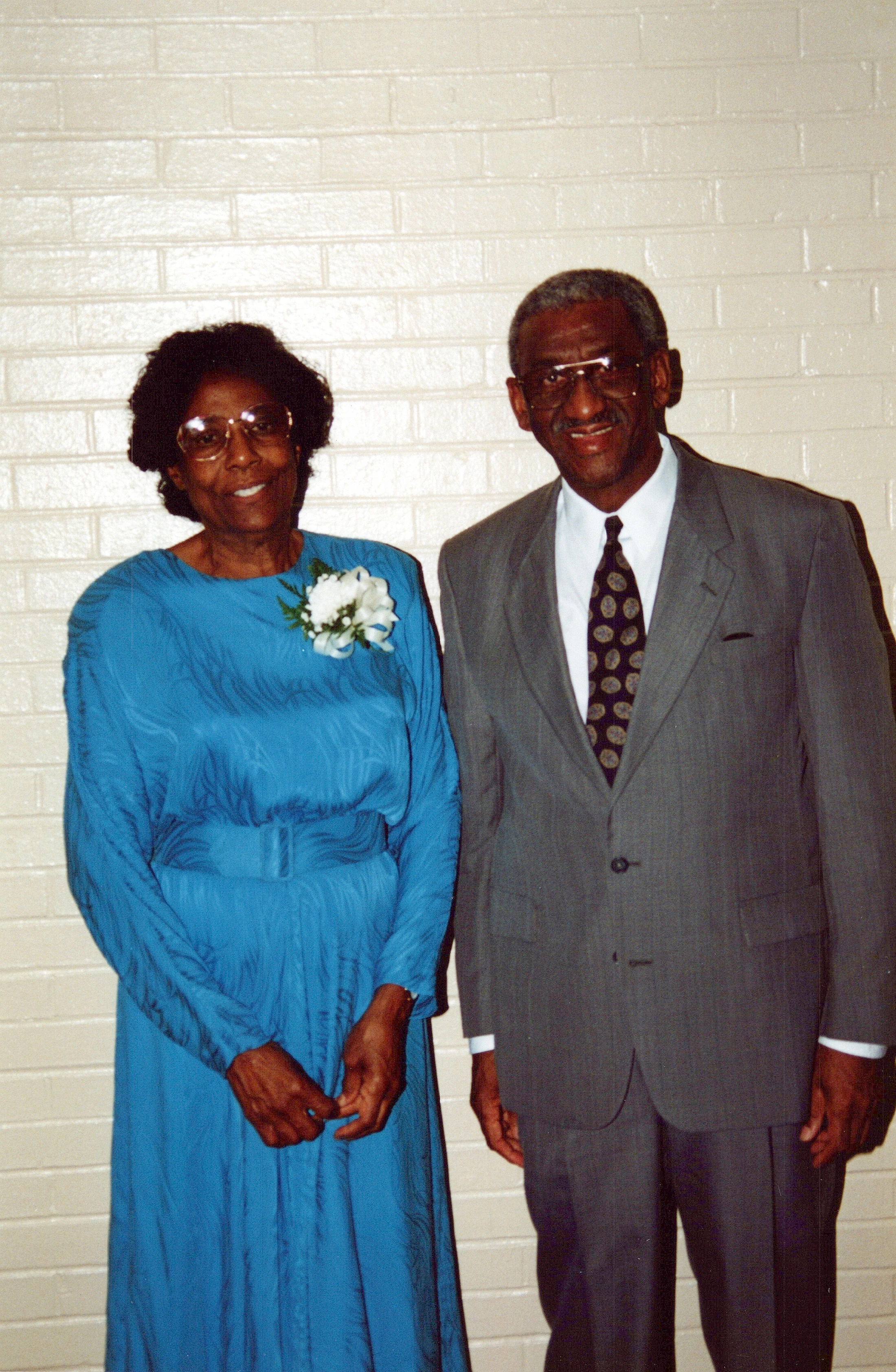 An older Black couple poses for a photo. The woman wears a blue dress with a corsage, and the man wears a gray suit. Both are smiling at the camera.