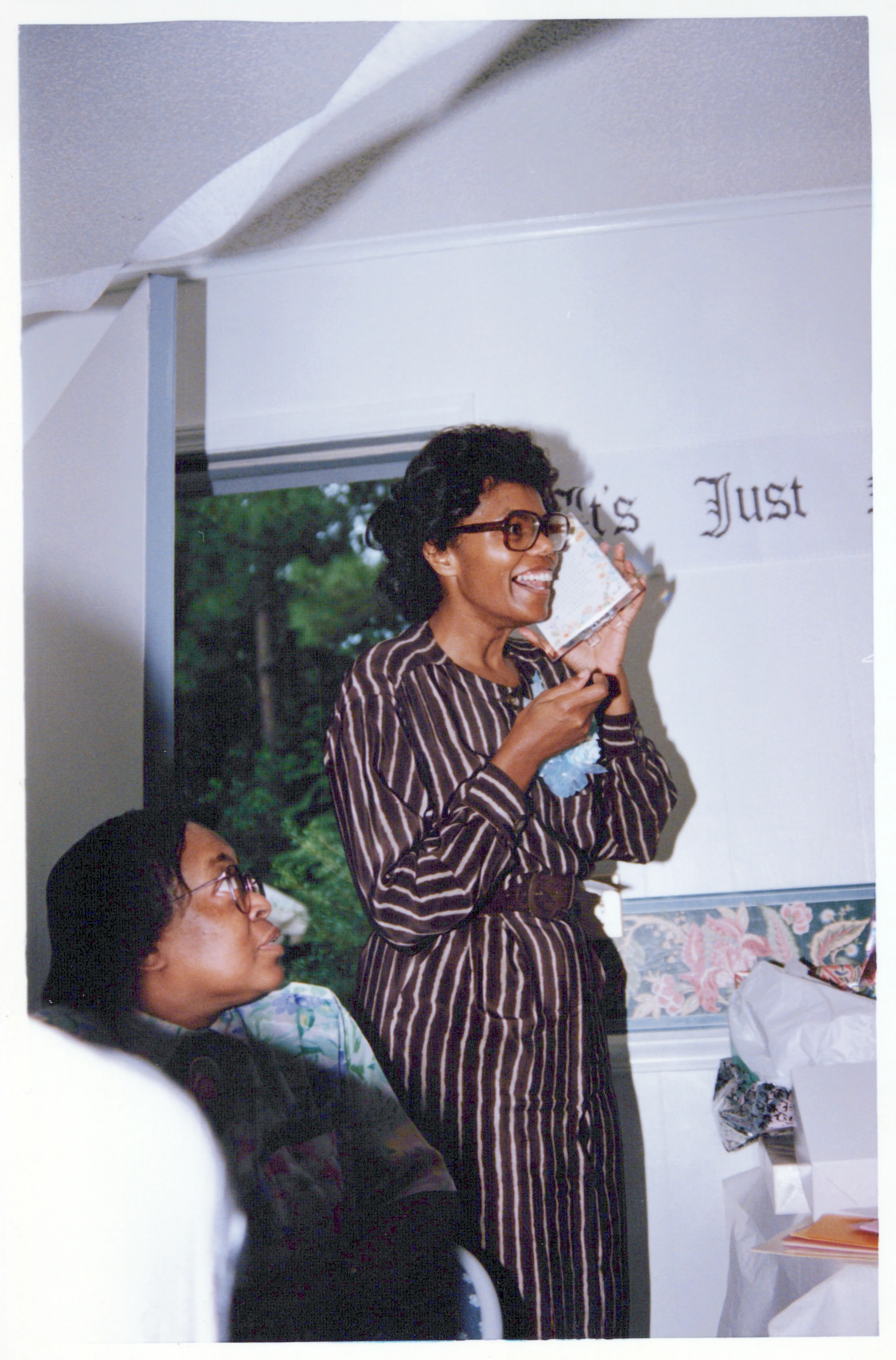A vintage photo of a Black woman in a striped dress and glasses, smiling with excitement as she opens a gift. Another woman in glasses watches her.