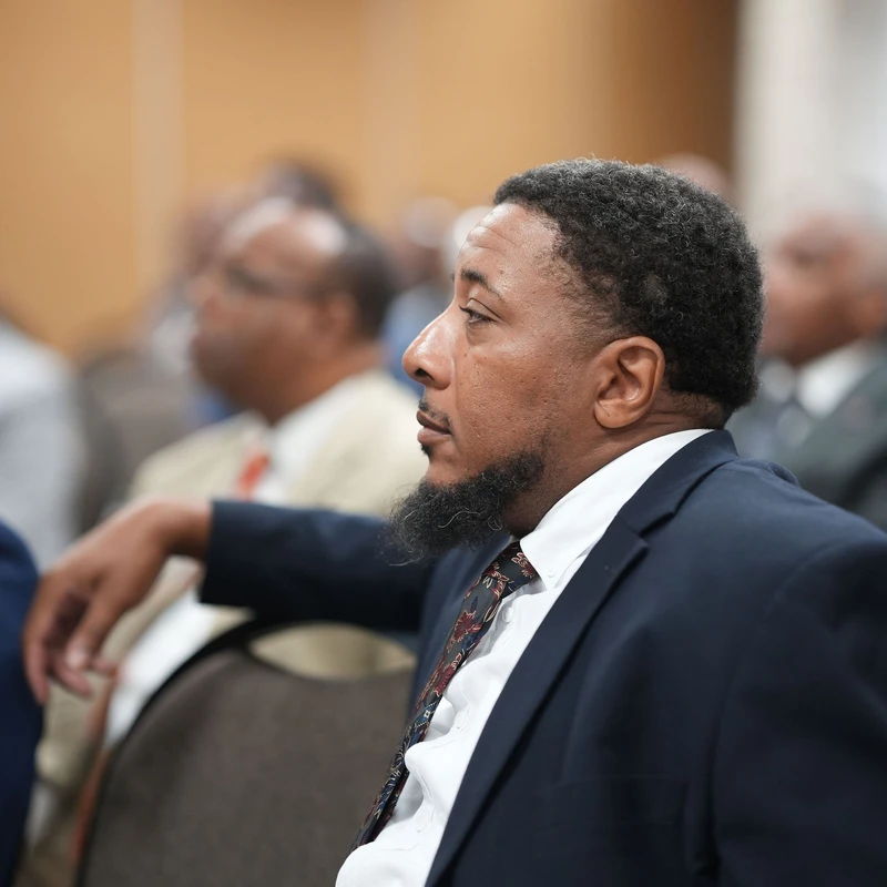 A man with a beard, wearing a dark suit and patterned tie, sits in profile in an audience, listening with a focused expression.
