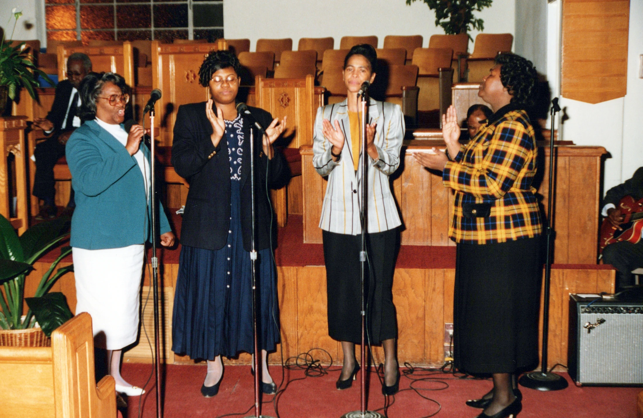 Four Black women stand on a carpeted stage in a church, singing and clapping into microphones. They are dressed up, and wooden pews are in the background.