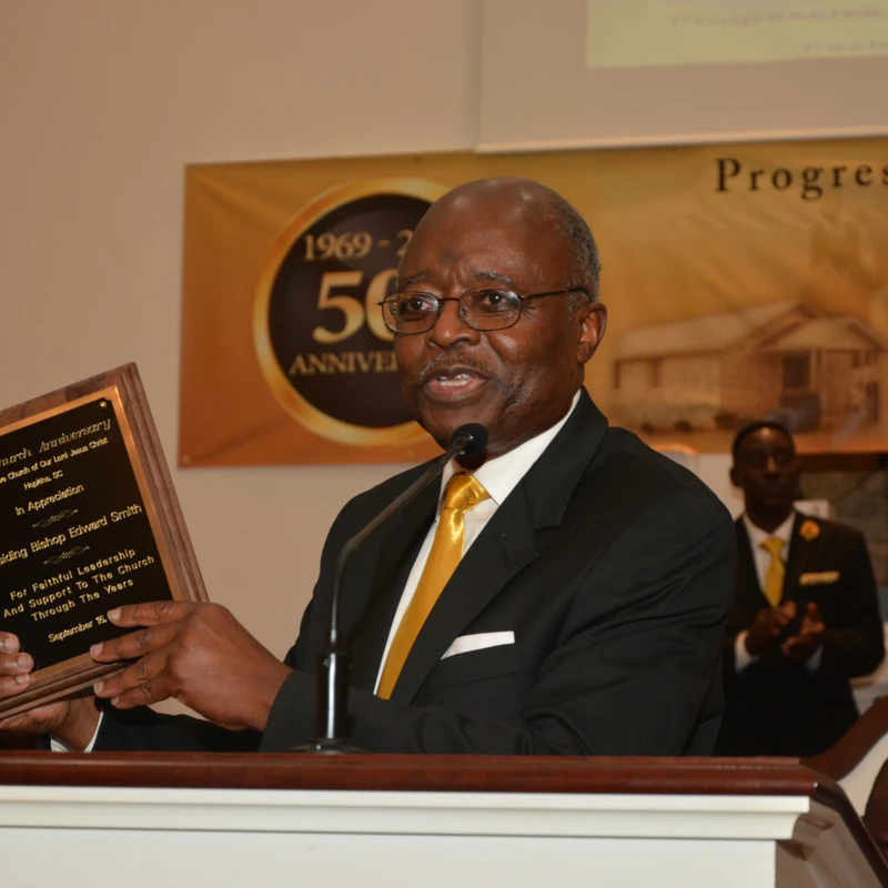 A man in a suit and glasses stands at a podium, holding up a wooden plaque and speaking into a microphone during a church anniversary event.