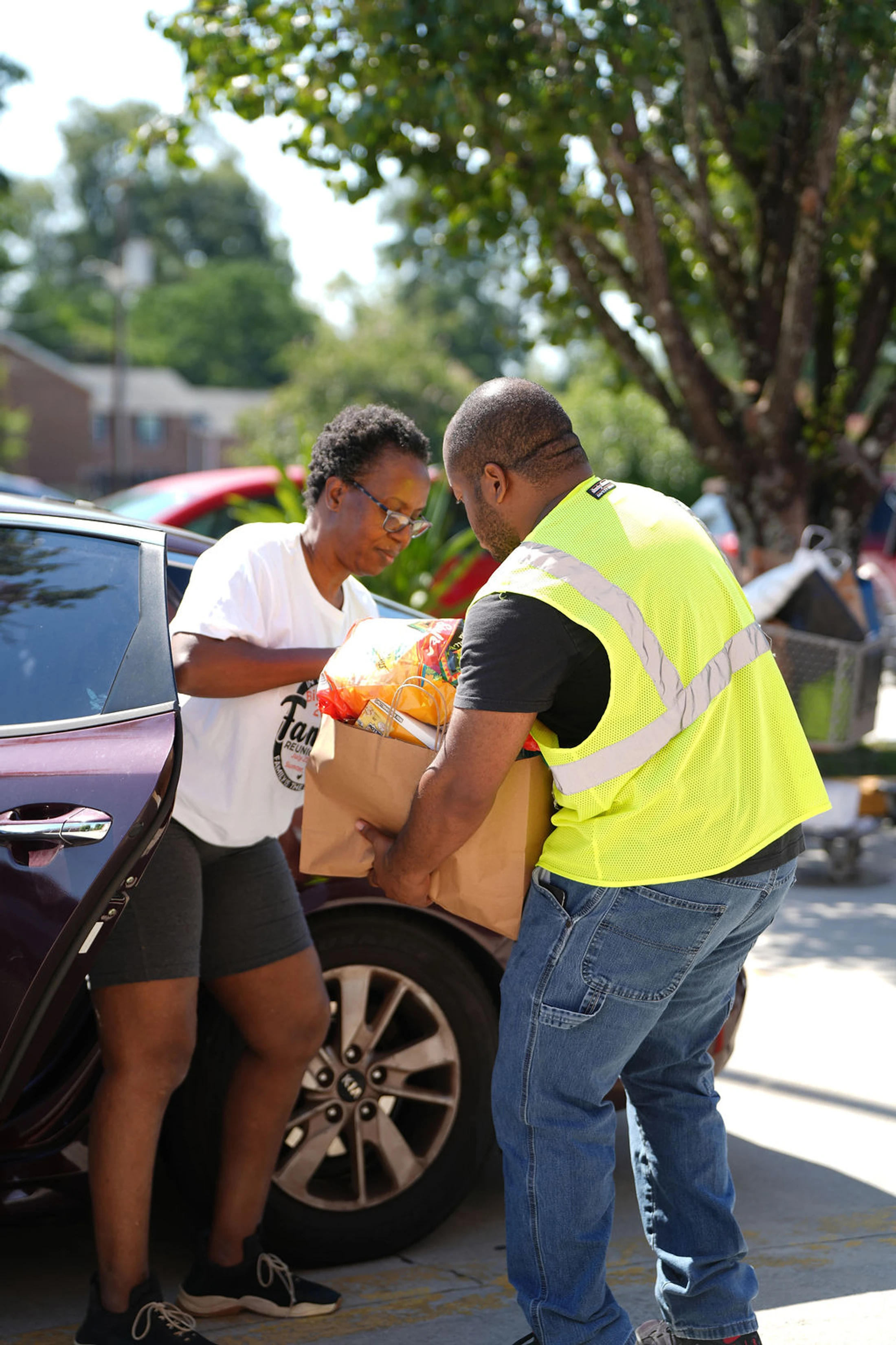 A male volunteer in a yellow safety vest helps a woman by loading a large paper bag full of groceries into her car at a community food drive.