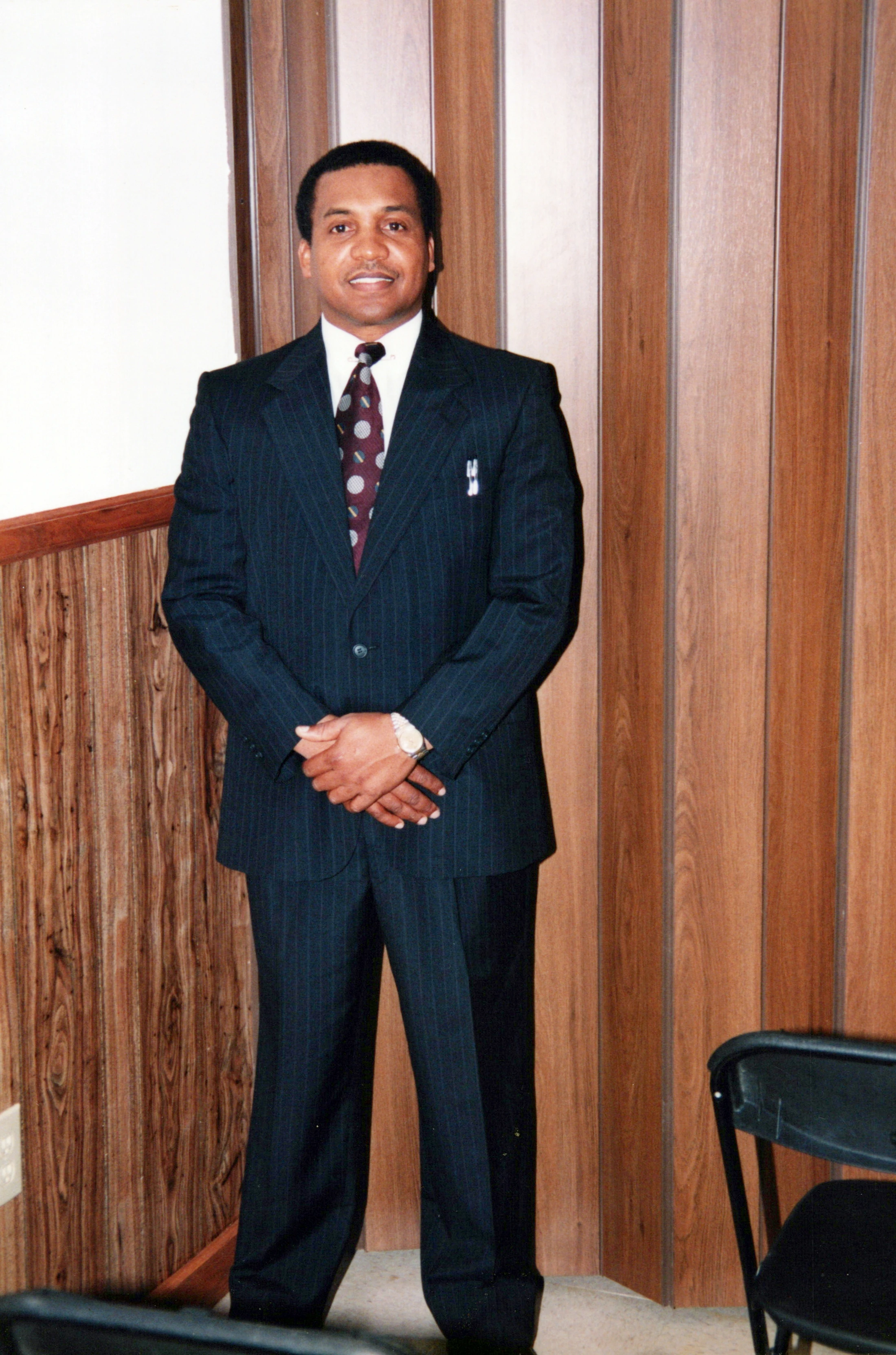 A full-length portrait of a smiling Black man in a dark pinstripe suit and patterned tie, standing with hands clasped in front of a wood-paneled wall.