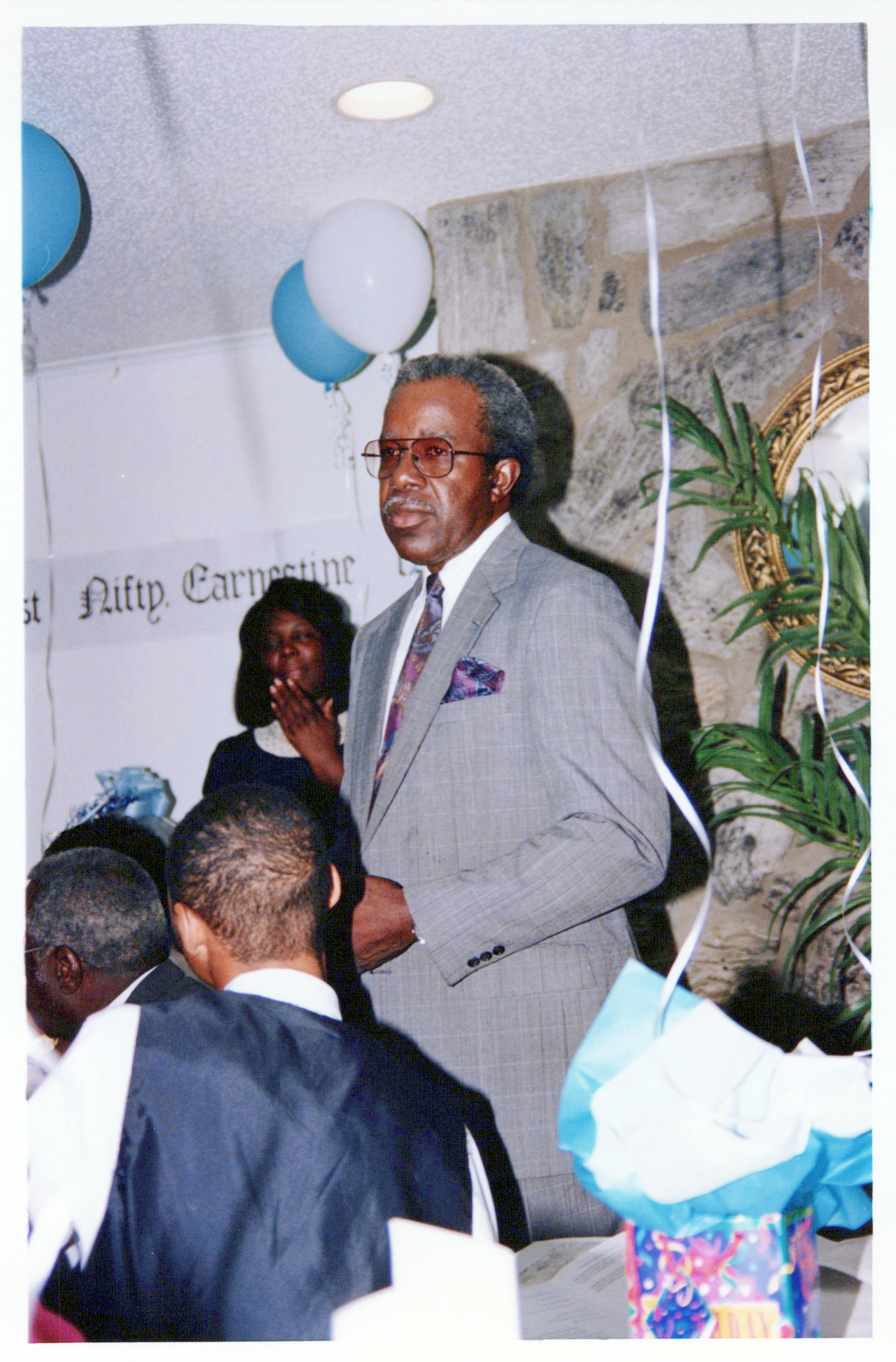 An older Black man with grey hair and glasses, wearing a light grey suit and tie, stands indoors at a party with balloons and other guests around him.