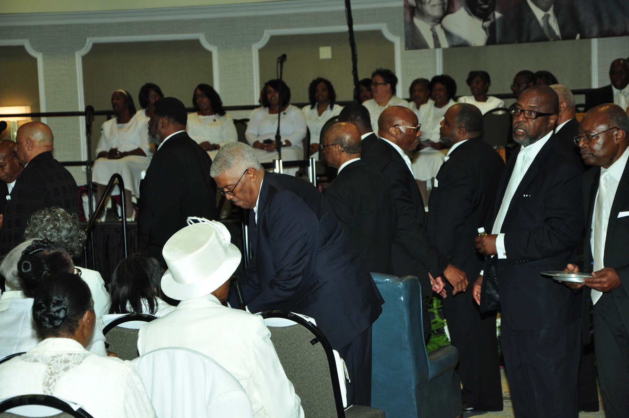 A congregation of African American men and women at a formal event. Men in dark suits stand in line, while women and a choir in white are seated.